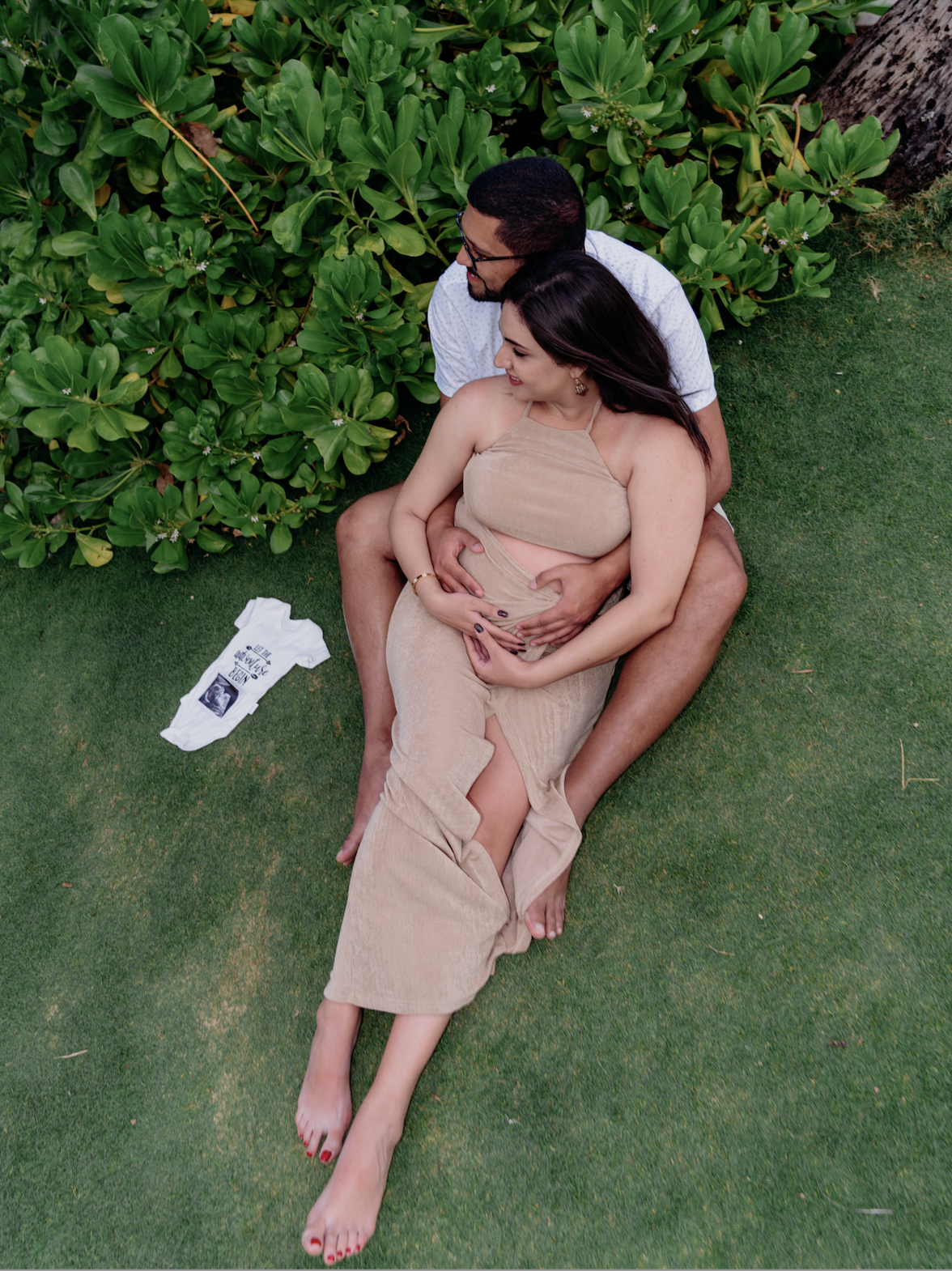 A pregnant woman and her partner sitting on the grass in a garden, with the woman cradling her belly and the man holding her. There is a white baby onesie on the grass nearby.
