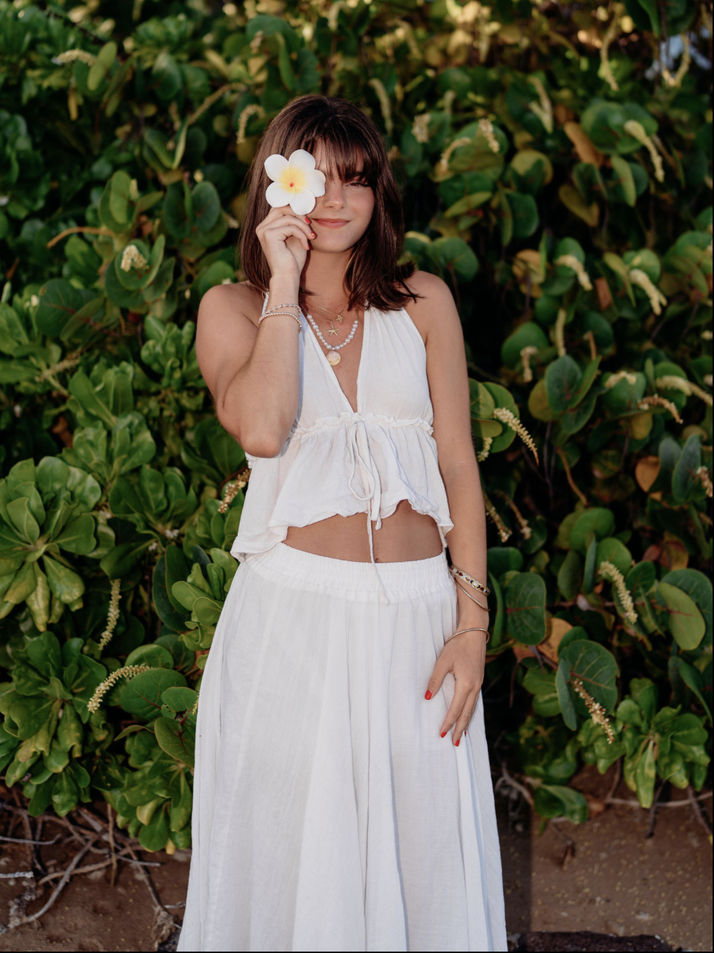 Young woman with brown hair in a white spaghetti strap top and white wide-leg pants, holding a white flower partially covering one eye, standing against green leafy bushes.