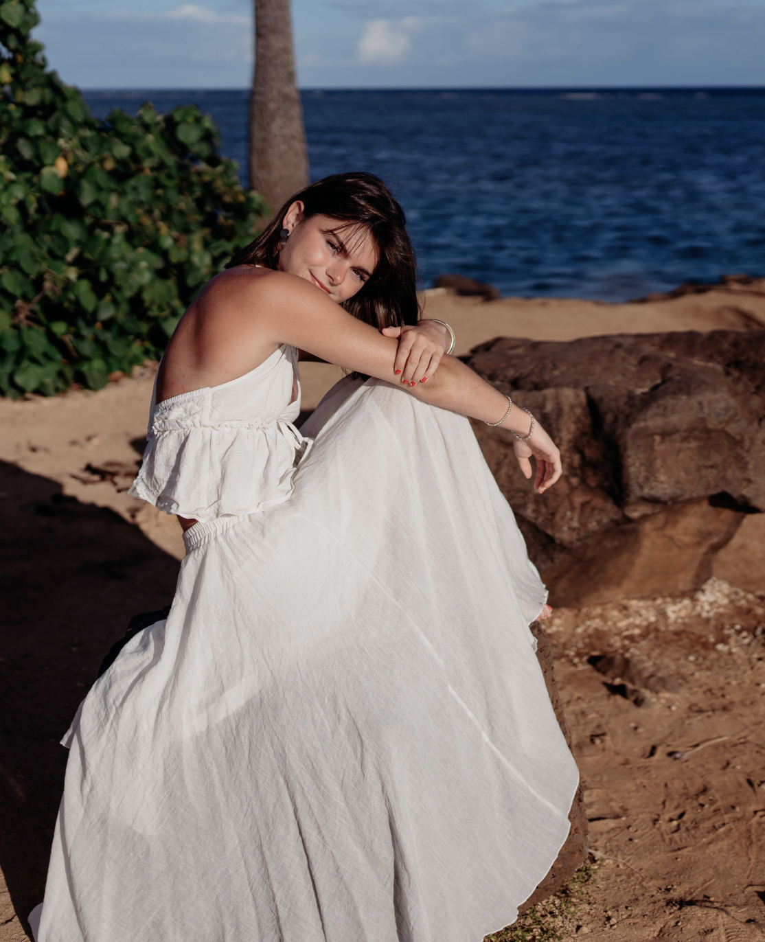 Woman sitting on rocks by the ocean, wearing a white dress, with her arms wrapped around her knee, smiling at the camera.