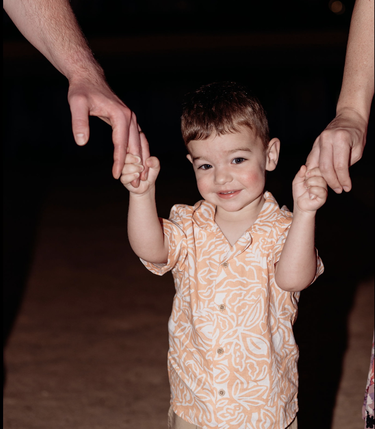 A young boy with short brown hair and a patterned orange and white shirt holding hands with two adults, one on each side, against a dark background.