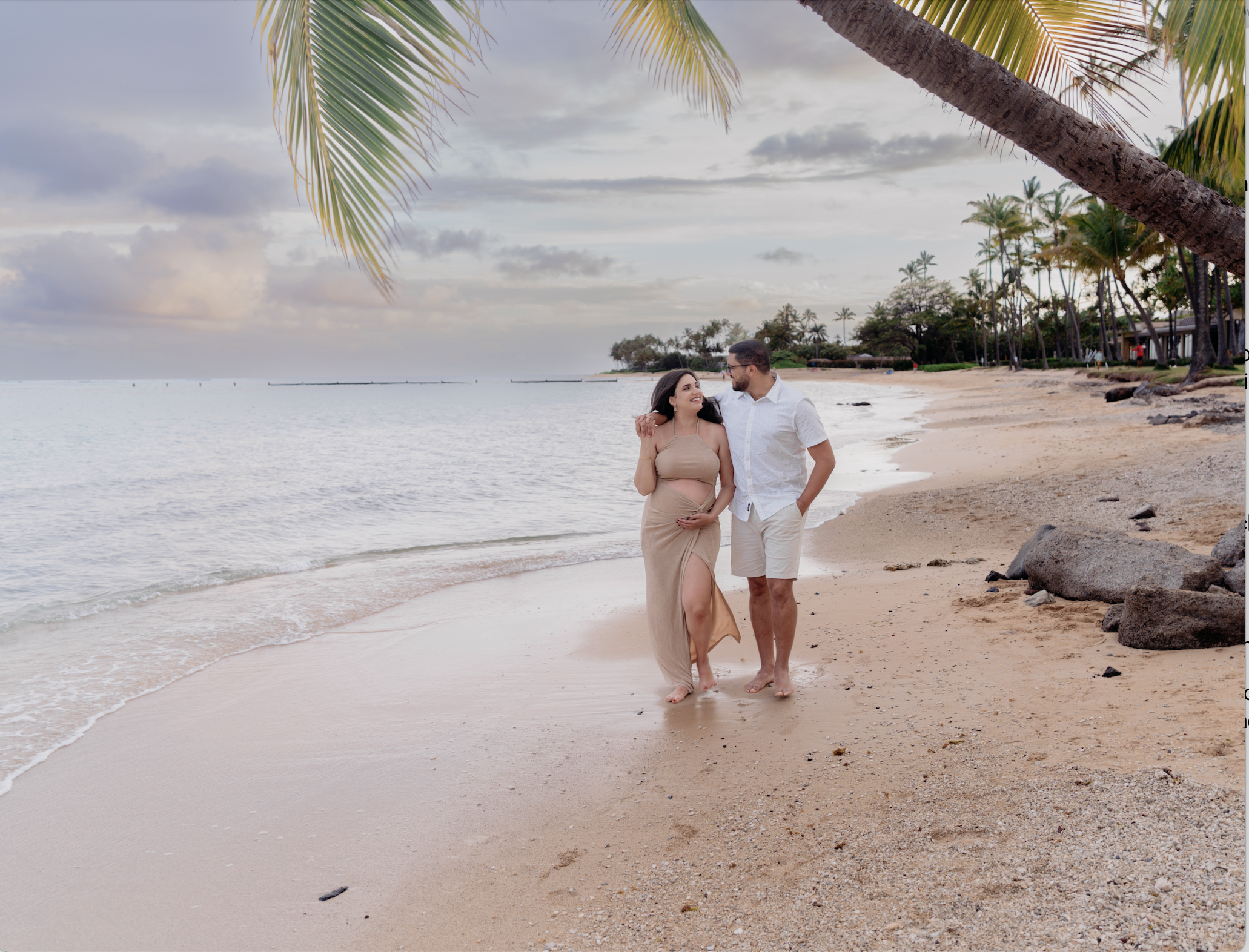 A pregnant woman and a man walking arm-in-arm along a tropical beach at sunset, with palm trees and a cloudy sky in the background.