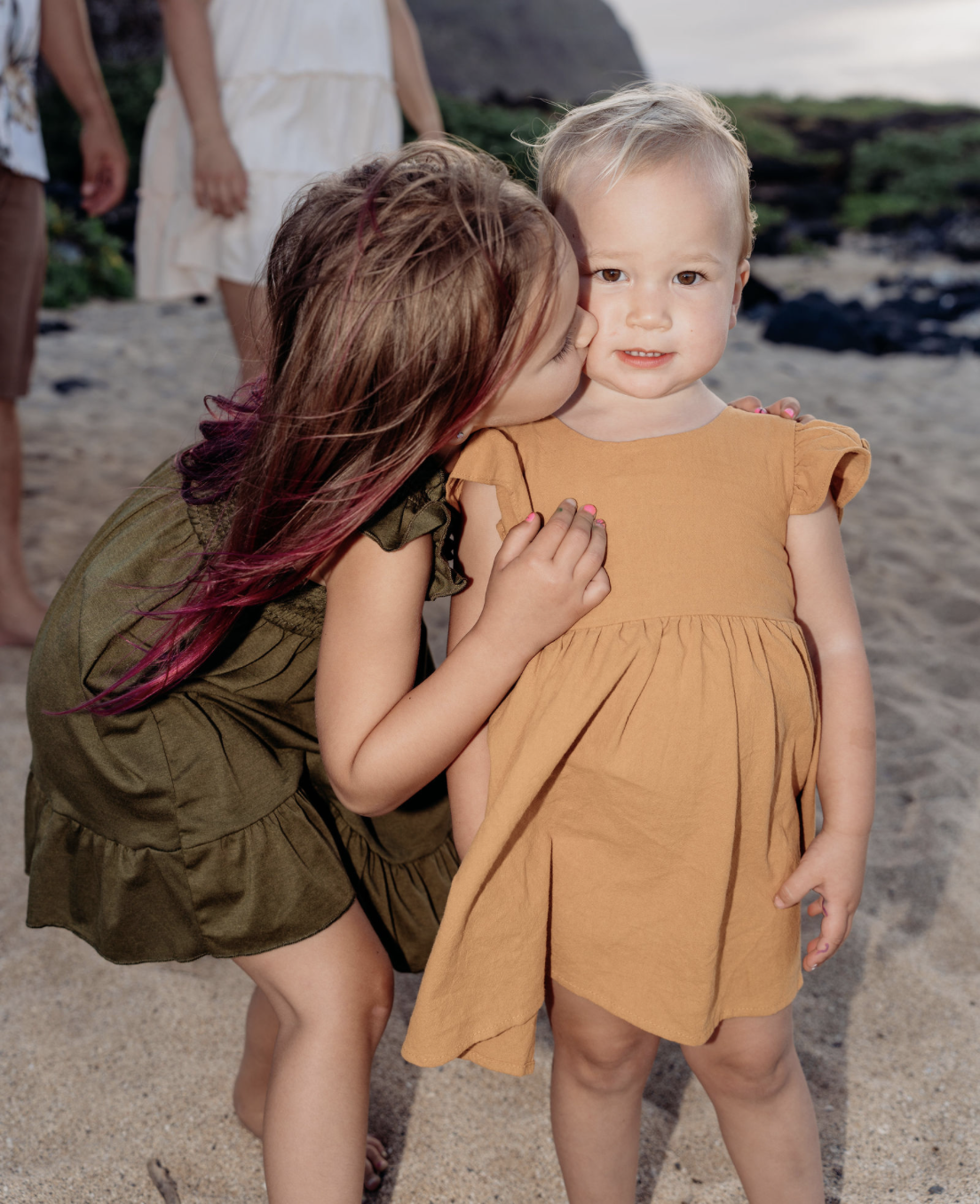A young girl with brown hair and pink highlights kissing a little girl with blonde hair on the cheek at the beach.