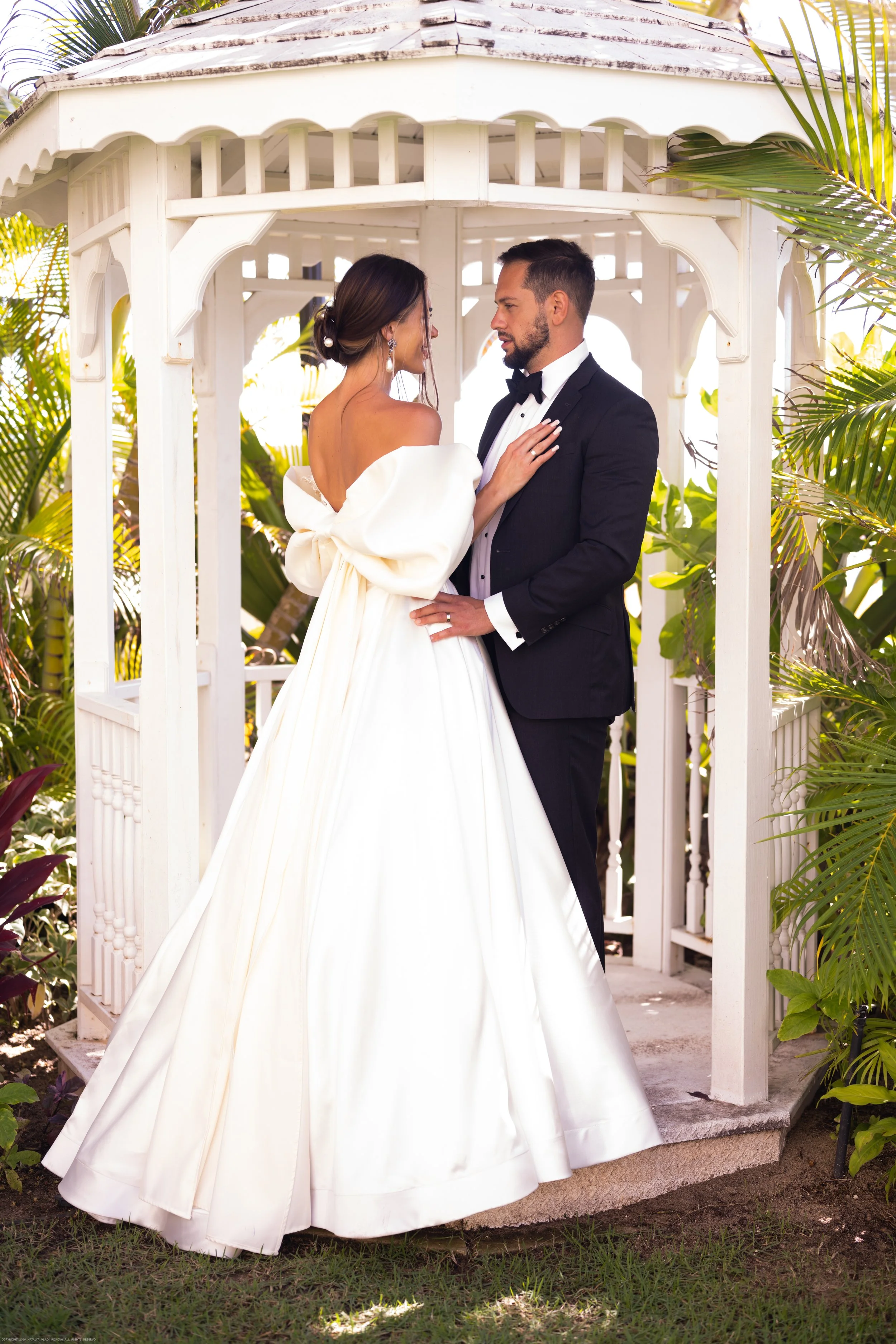 A bride and groom standing close together inside a white gazebo surrounded by tropical plants.