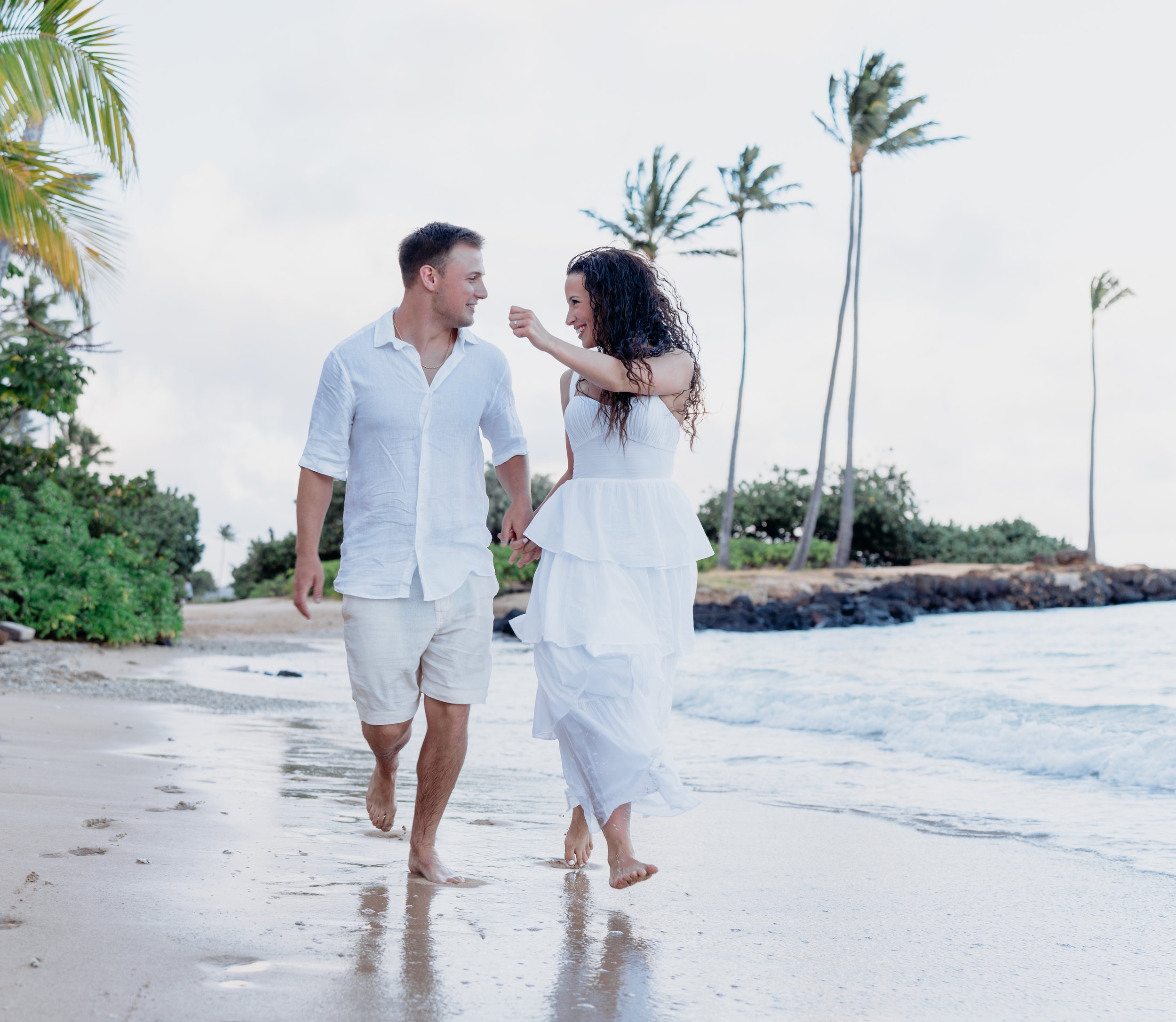 A couple dressed in white walking barefoot on a beach, holding hands, with palm trees and ocean in the background.