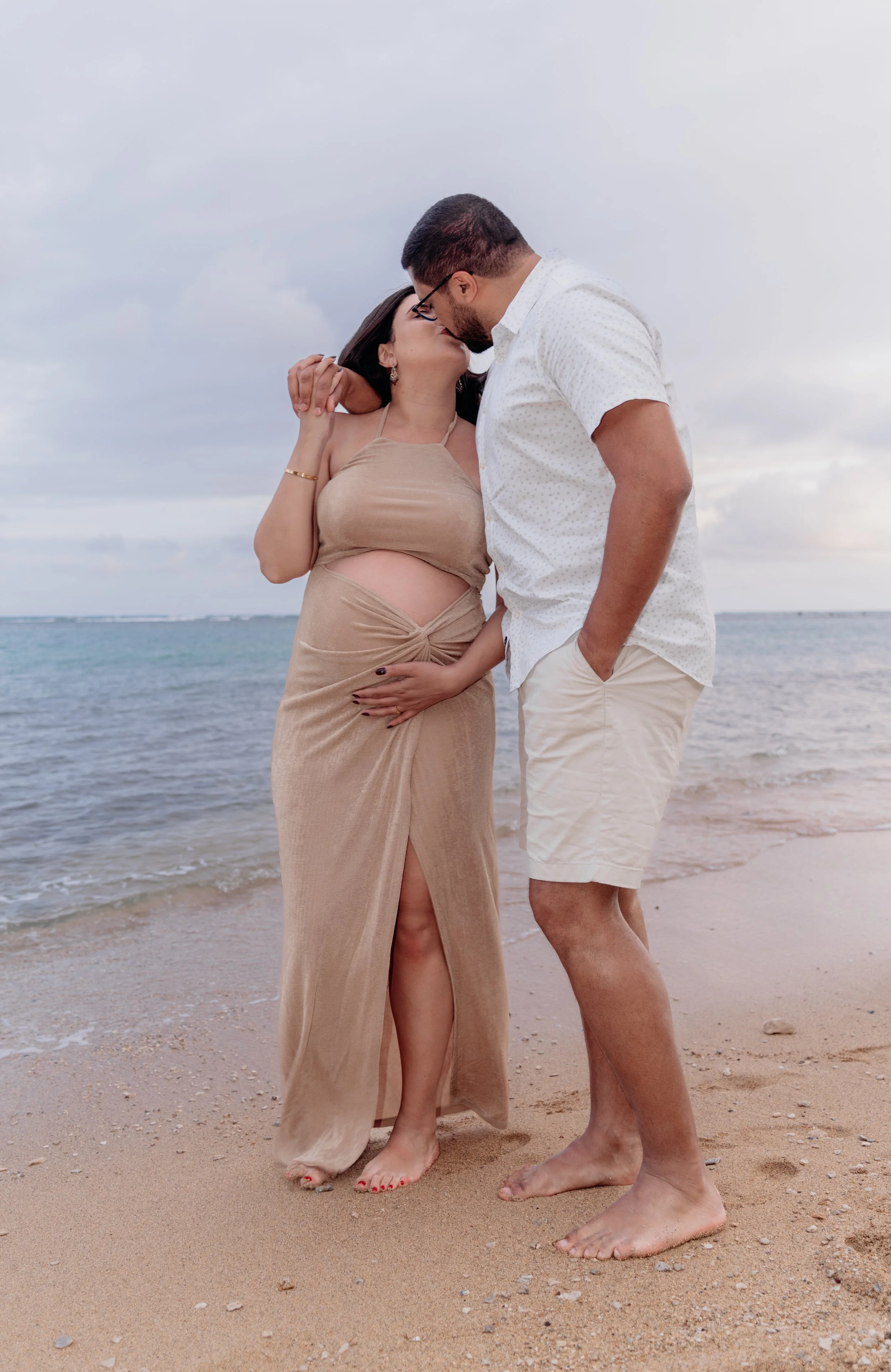 A couple sharing a kiss on the beach, with the woman pregnant, during a cloudy day.