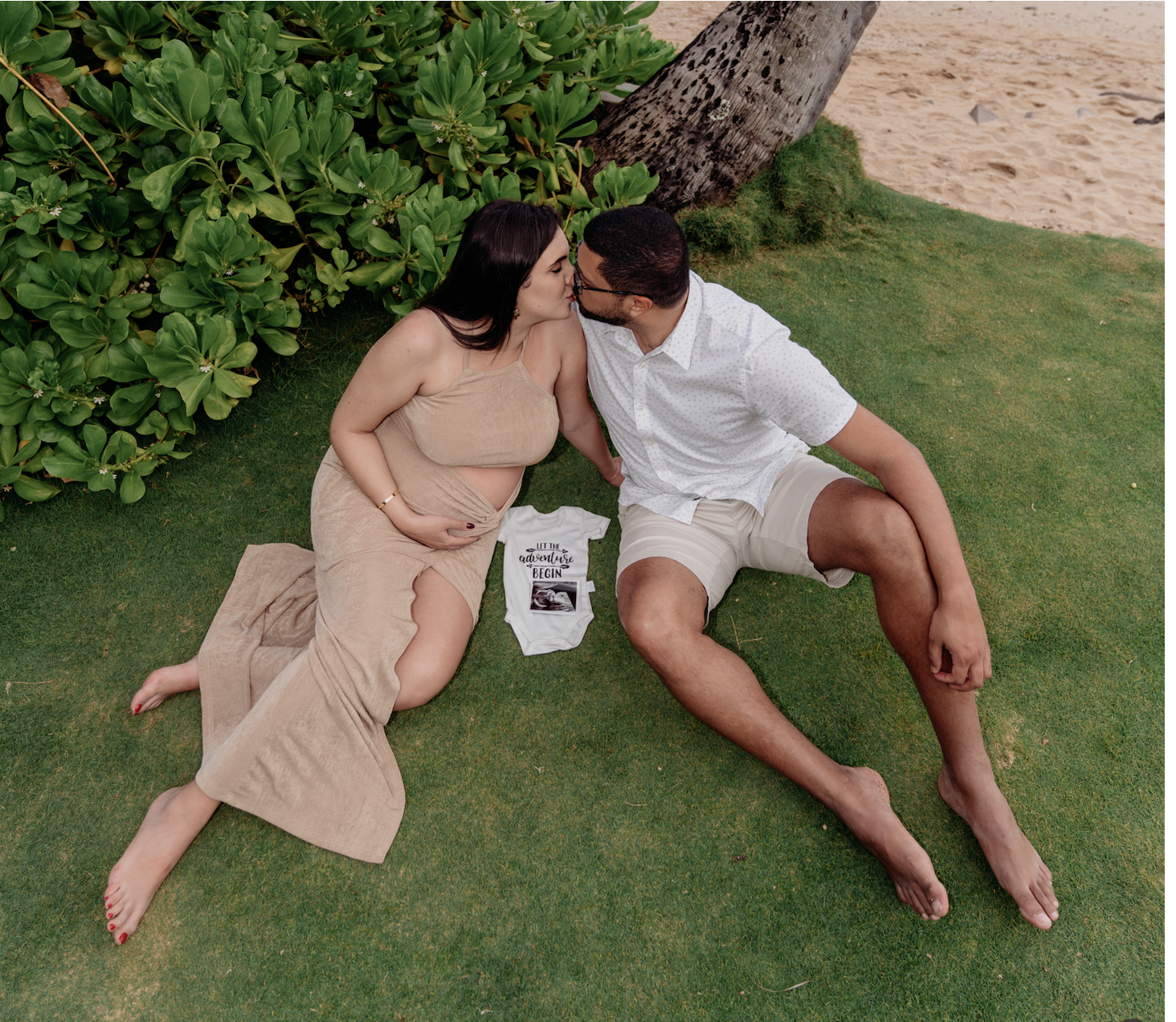 A pregnant woman and a man sitting on grass, leaning in to kiss, with a lush green plant and a tree in the background, and a sandy beach nearby.