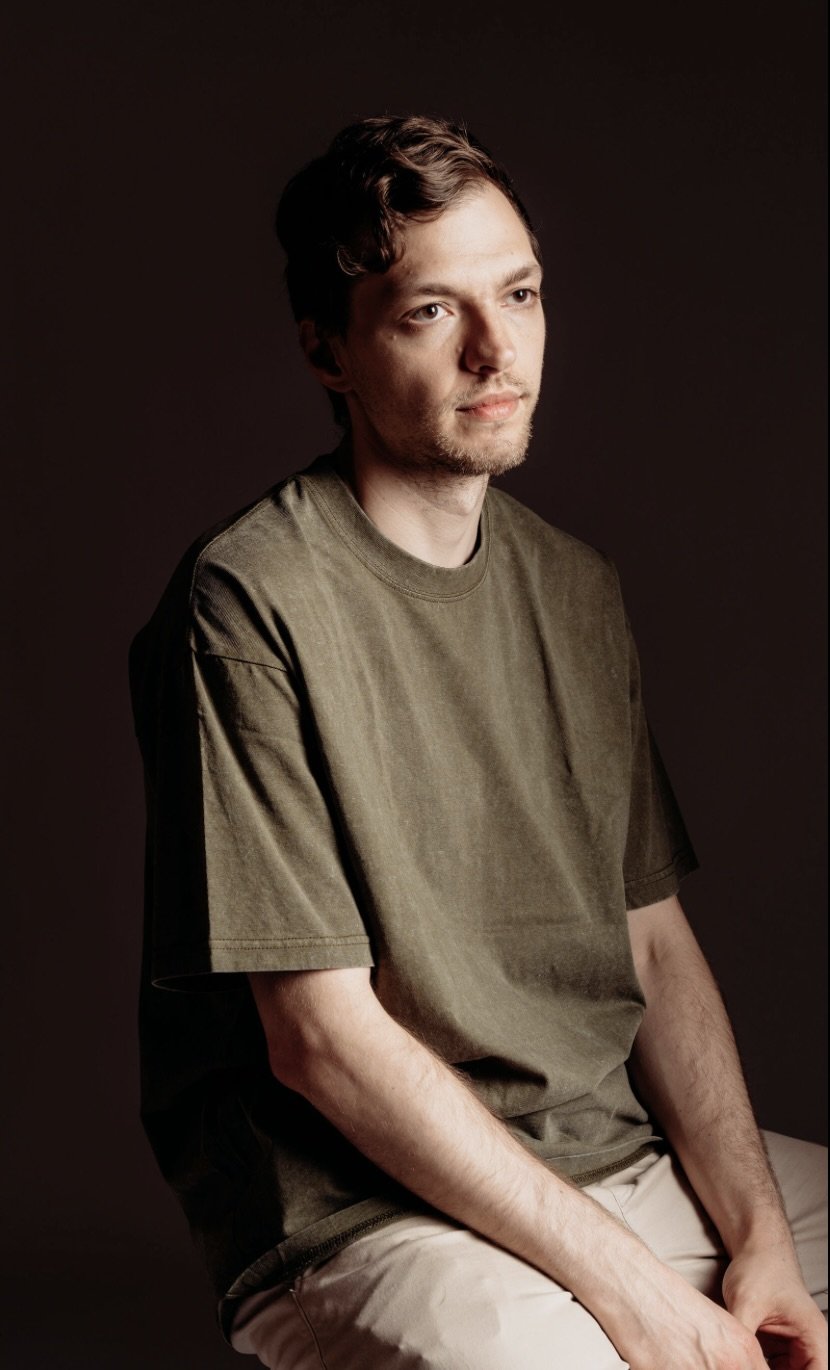 A young man with short brown hair and a light beard, wearing a khaki t-shirt and beige pants, sitting against a dark background, looking slightly to the side.