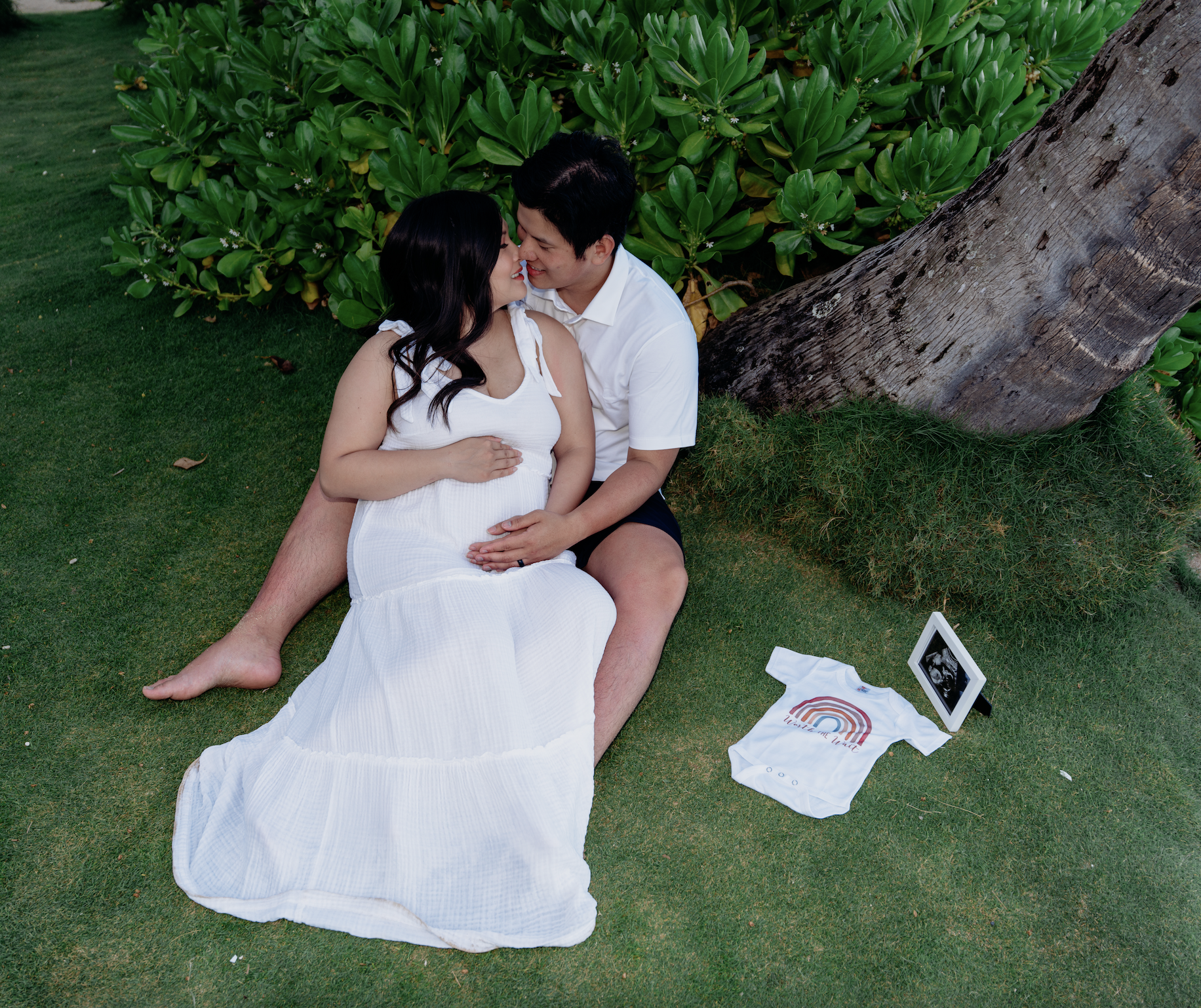 A pregnant woman and a man, sitting on grass next to a tree, are about to kiss. The woman is in a white dress and the man is in shorts and a white shirt. There is a white baby onesie and a tablet on the grass nearby.