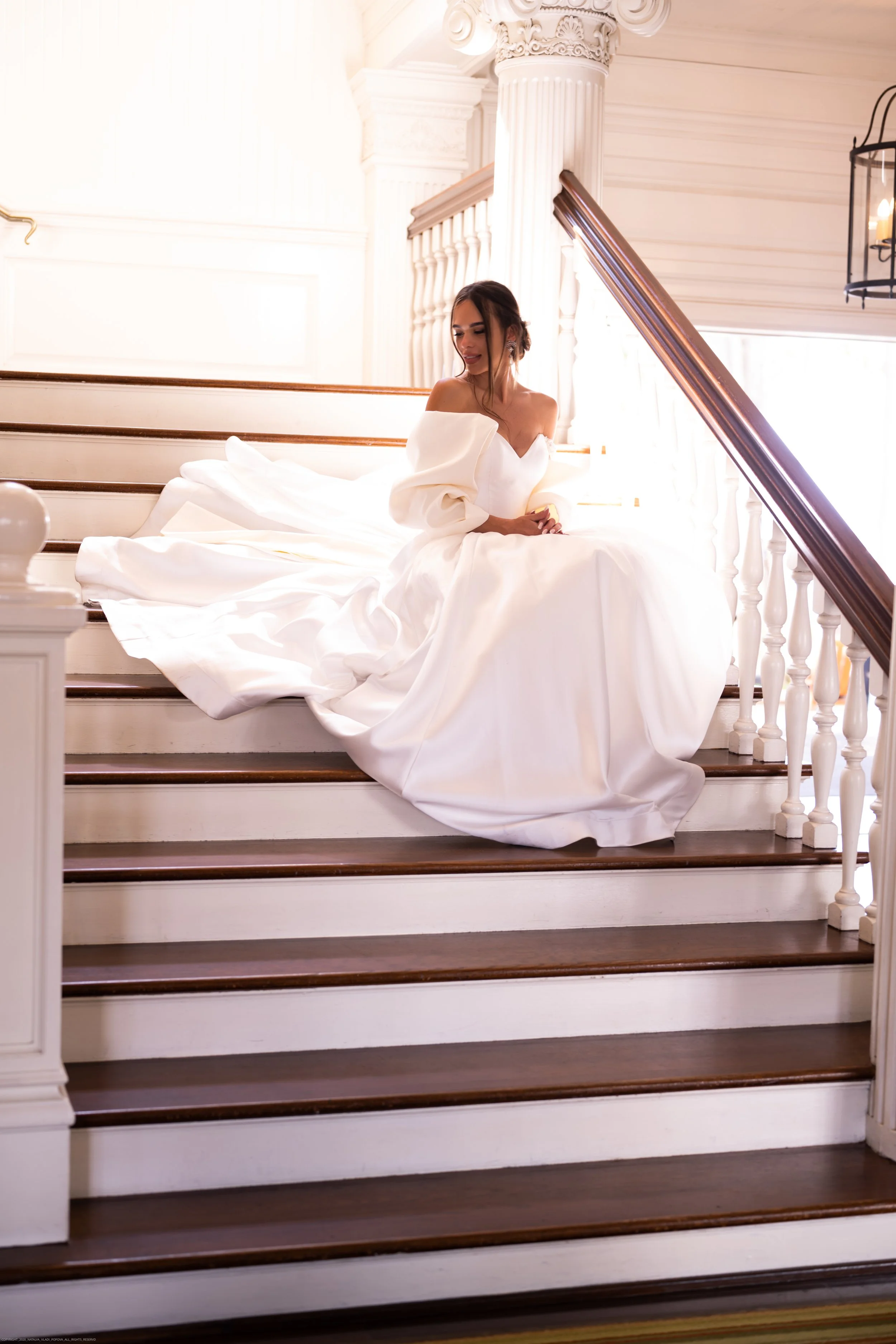 A woman in a white off-shoulder gown sitting on wooden stairs inside a bright, elegant room with white columns and decorative trim.