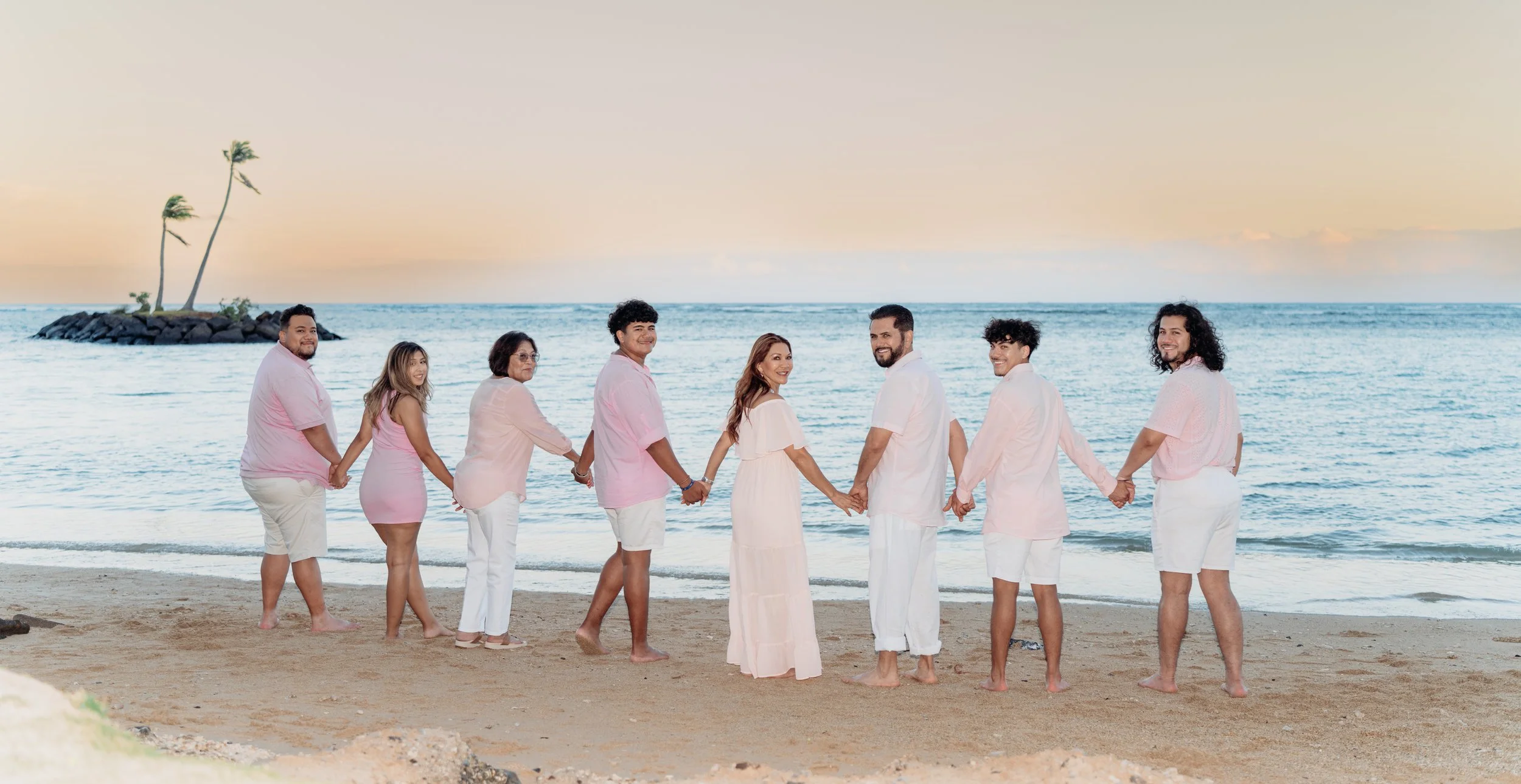 Group of nine people holding hands on a beach at sunset, wearing light-colored clothing, with the ocean and a small rocky island with palm trees in the background.