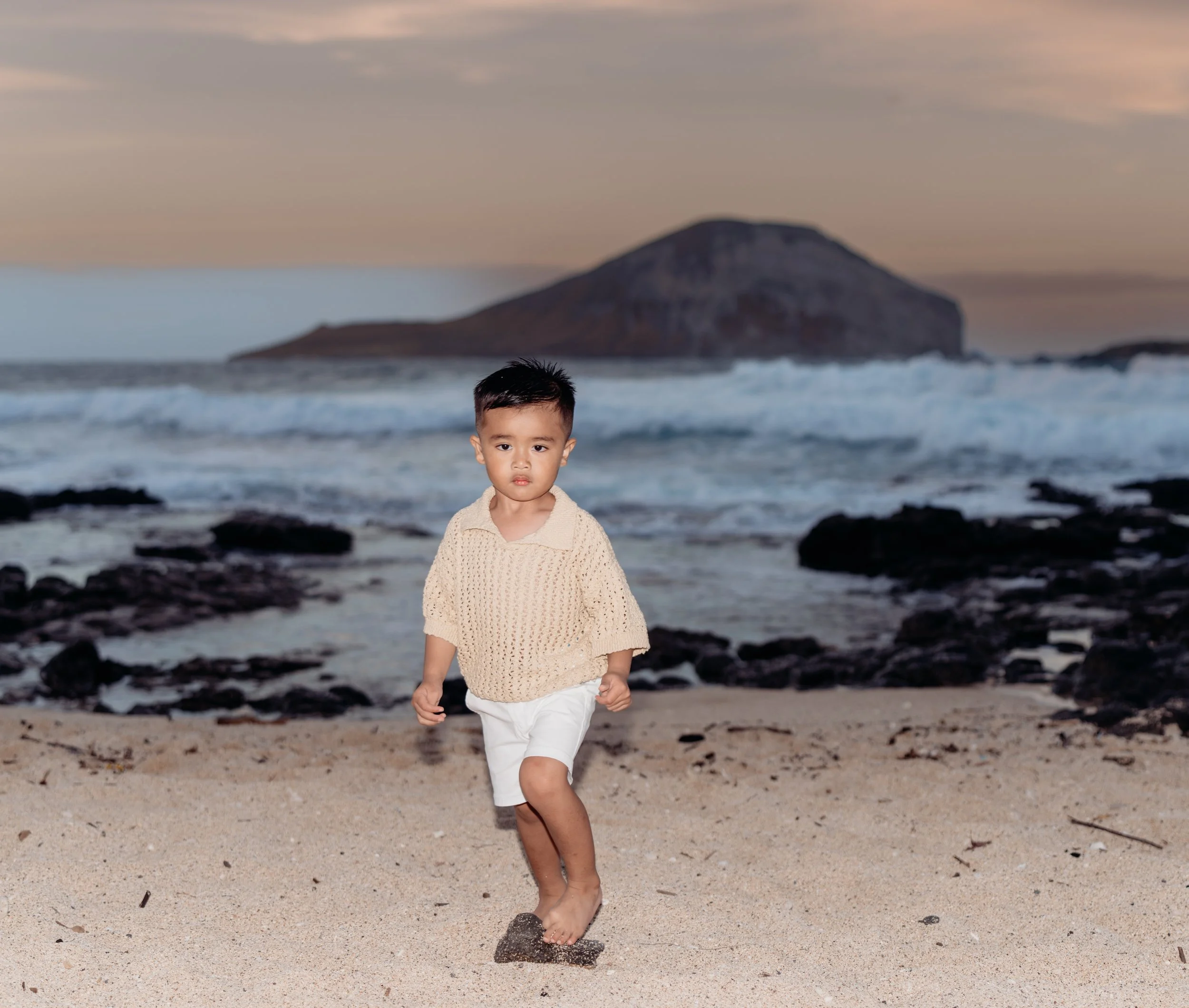 Young boy walking barefoot on a sandy beach with rocks, ocean waves, and a large island or mountain in the background during sunset.