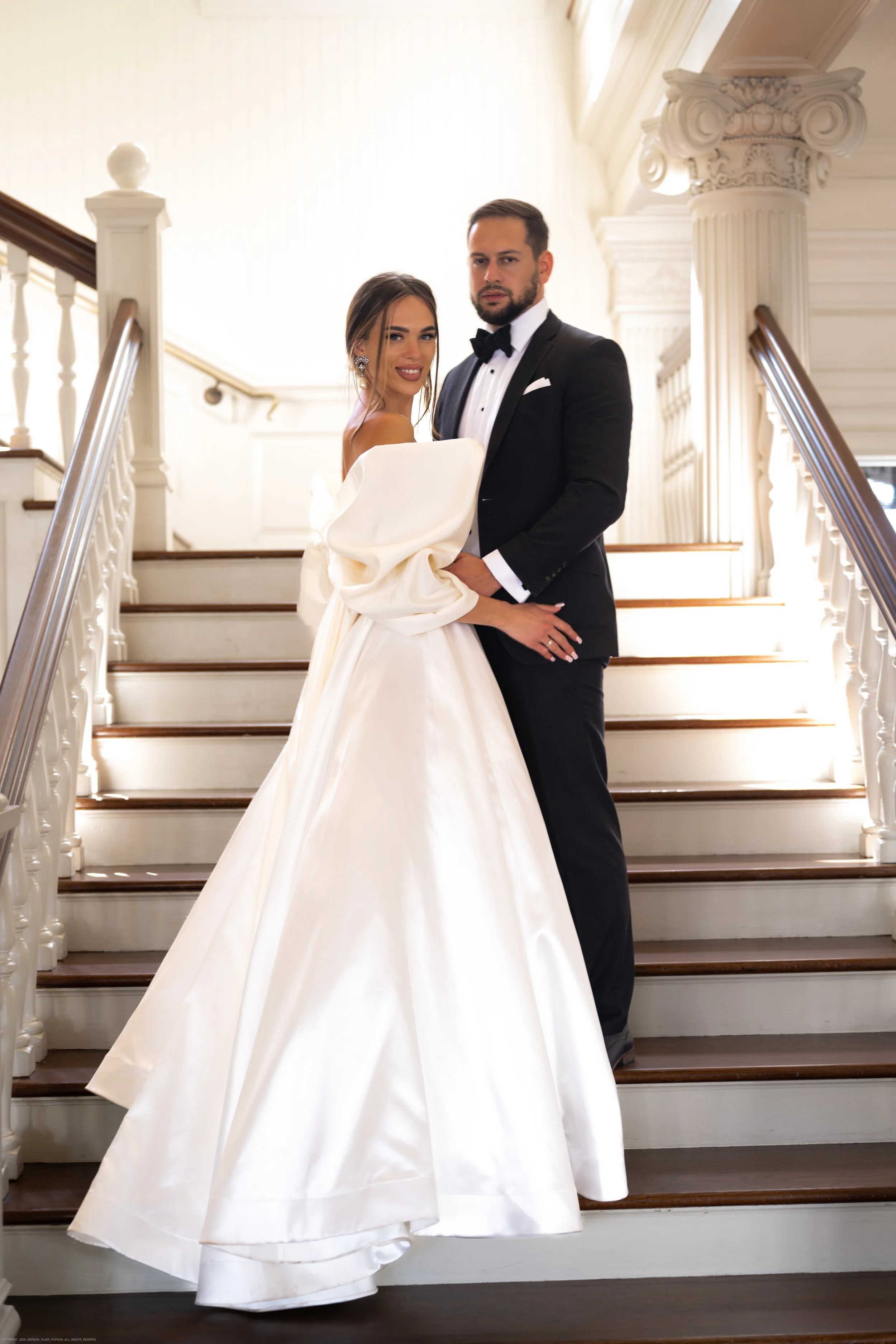 A bride and groom on a staircase in a well-lit, elegant venue. The bride is wearing a white wedding gown with puffed sleeves, and the groom is dressed in a black tuxedo with a bow tie. They are holding each other and looking at the camera.