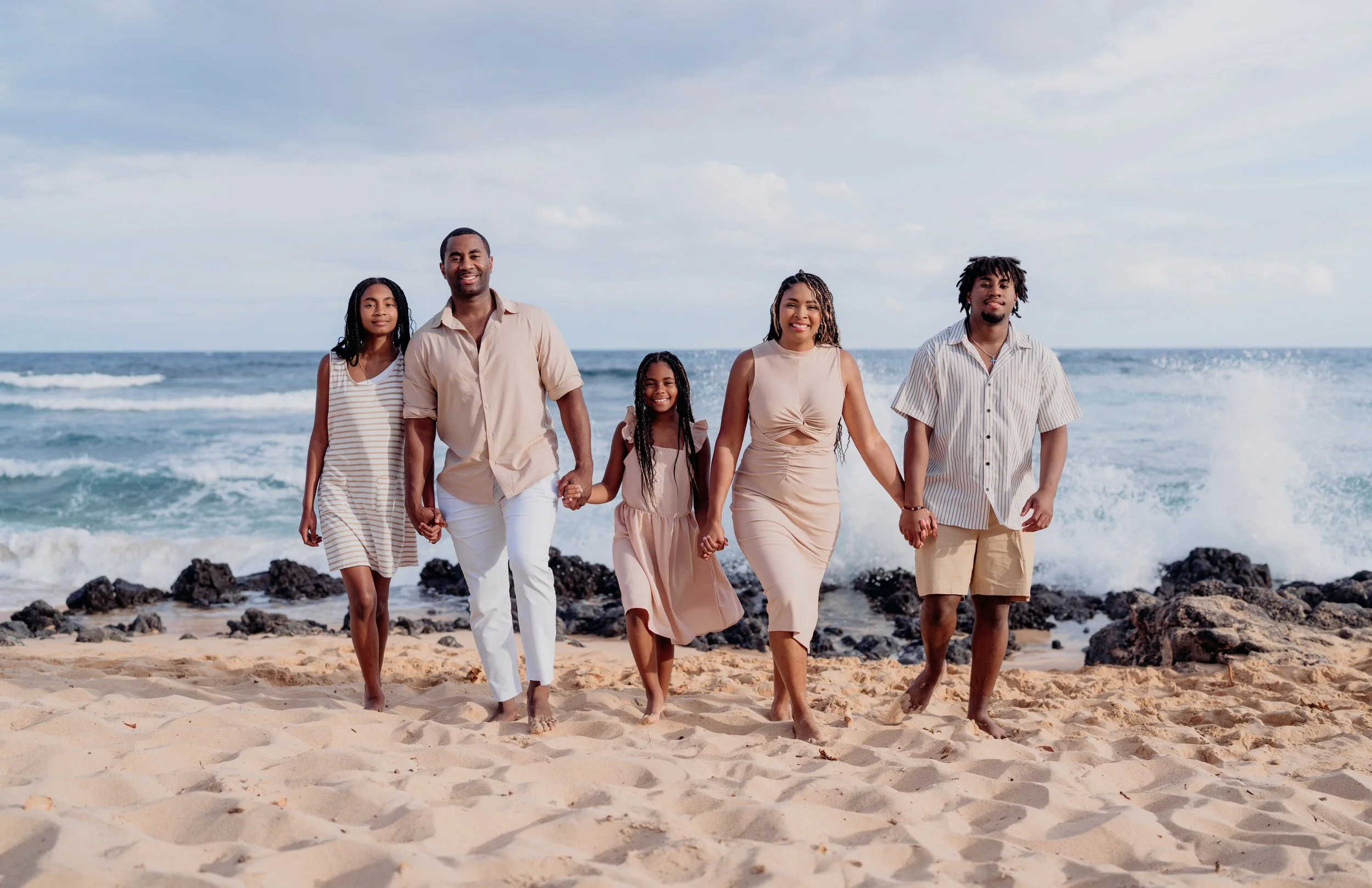 A family walking on a sandy beach with waves and rocks in the background, holding hands and smiling.