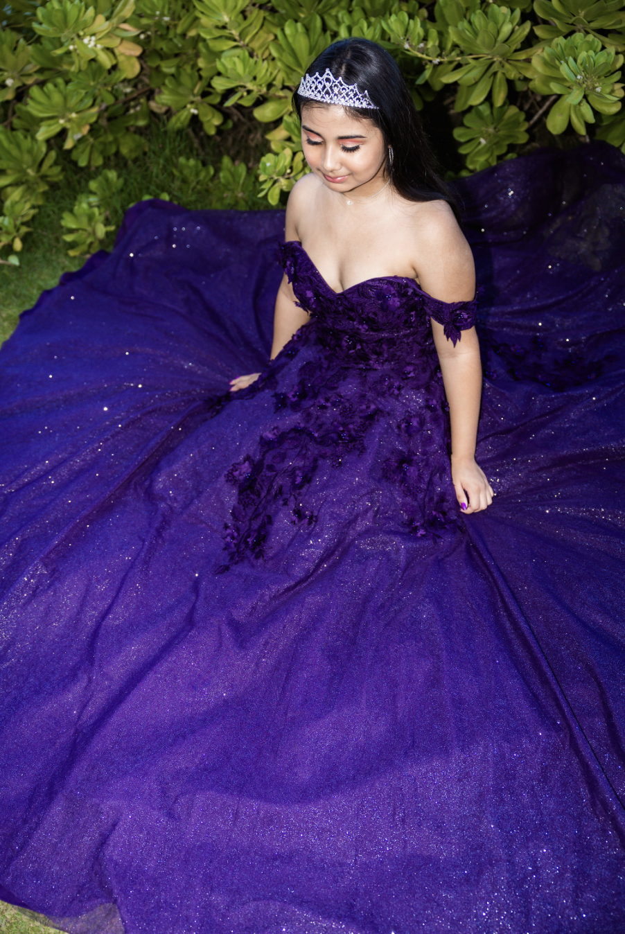 Young woman wearing a purple gown with off-the-shoulder sleeves, lying on the grass near green bushes, with a silver tiara on her head.