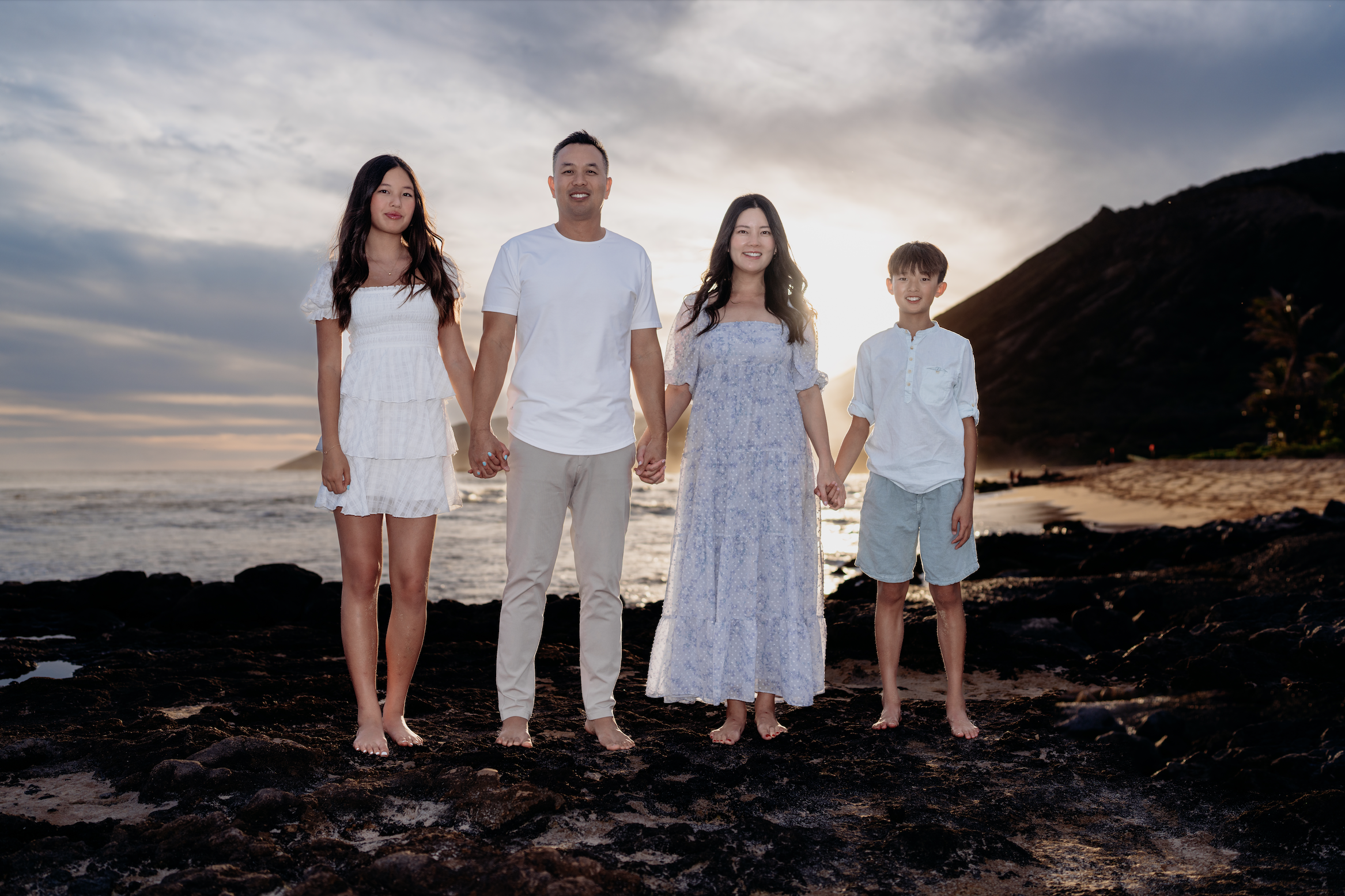 Family of four standing on a rocky beach at sunset, holding hands, with ocean and mountains in the background, dressed in white and light-colored clothing.