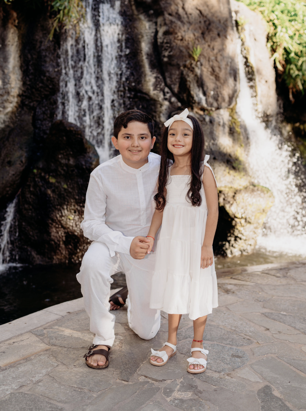 A boy and girl holding hands and smiling in front of a waterfall. The boy is kneeling and wearing a white outfit with sandals, while the girl is standing in a white dress with a headband and sandals.
