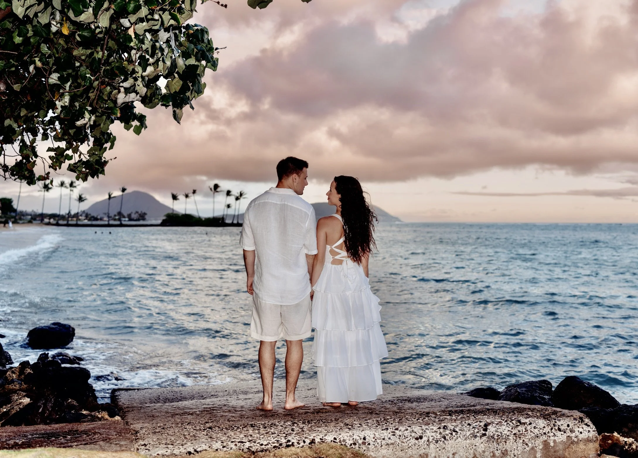 A couple stands on a concrete platform at the beach, facing each other with the ocean and a mountain in the background during sunset. The woman is wearing a white dress with cutouts on the back, and the man is dressed in a light-colored shirt and sho