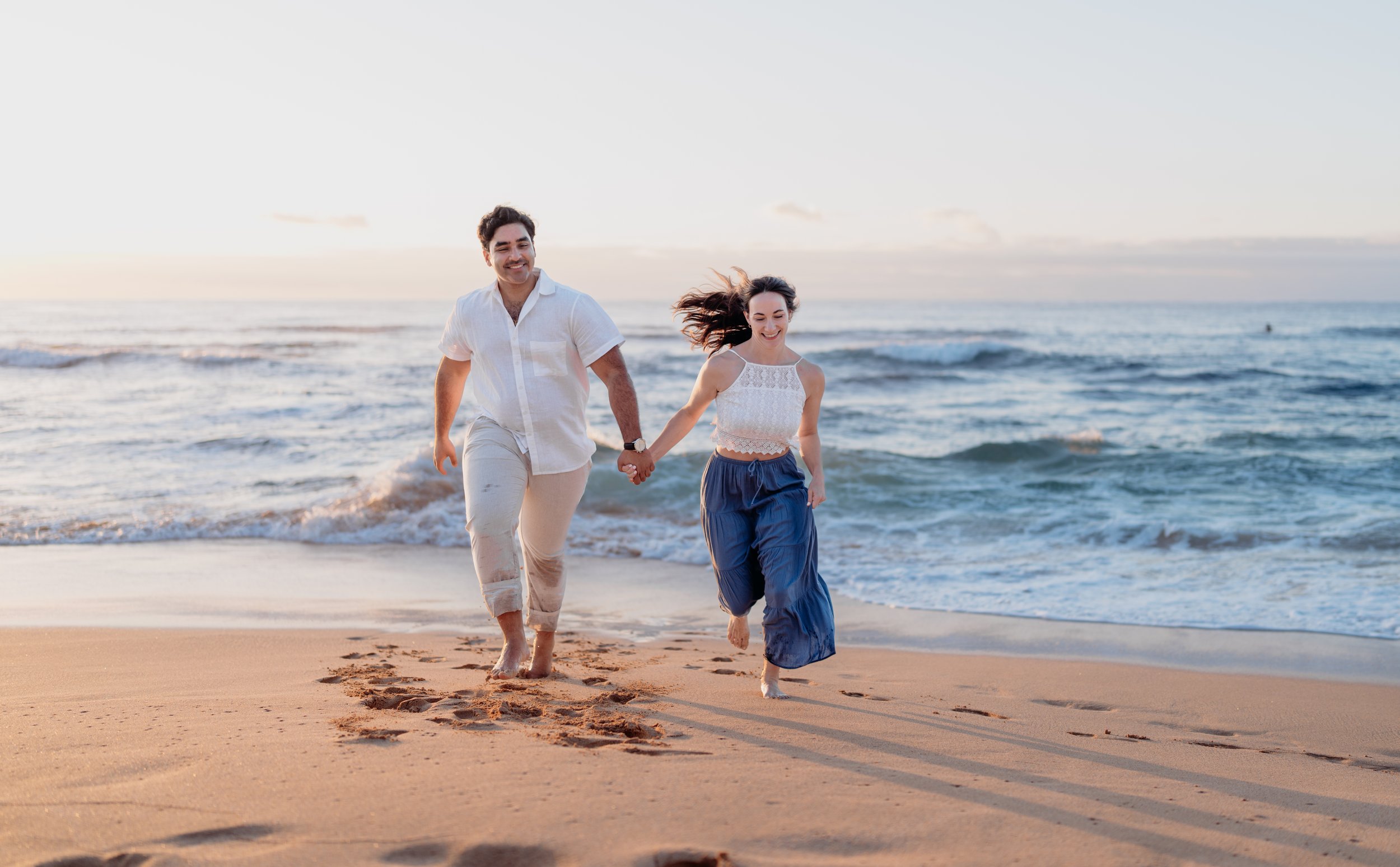 A young man and woman holding hands and running on the beach during sunset.