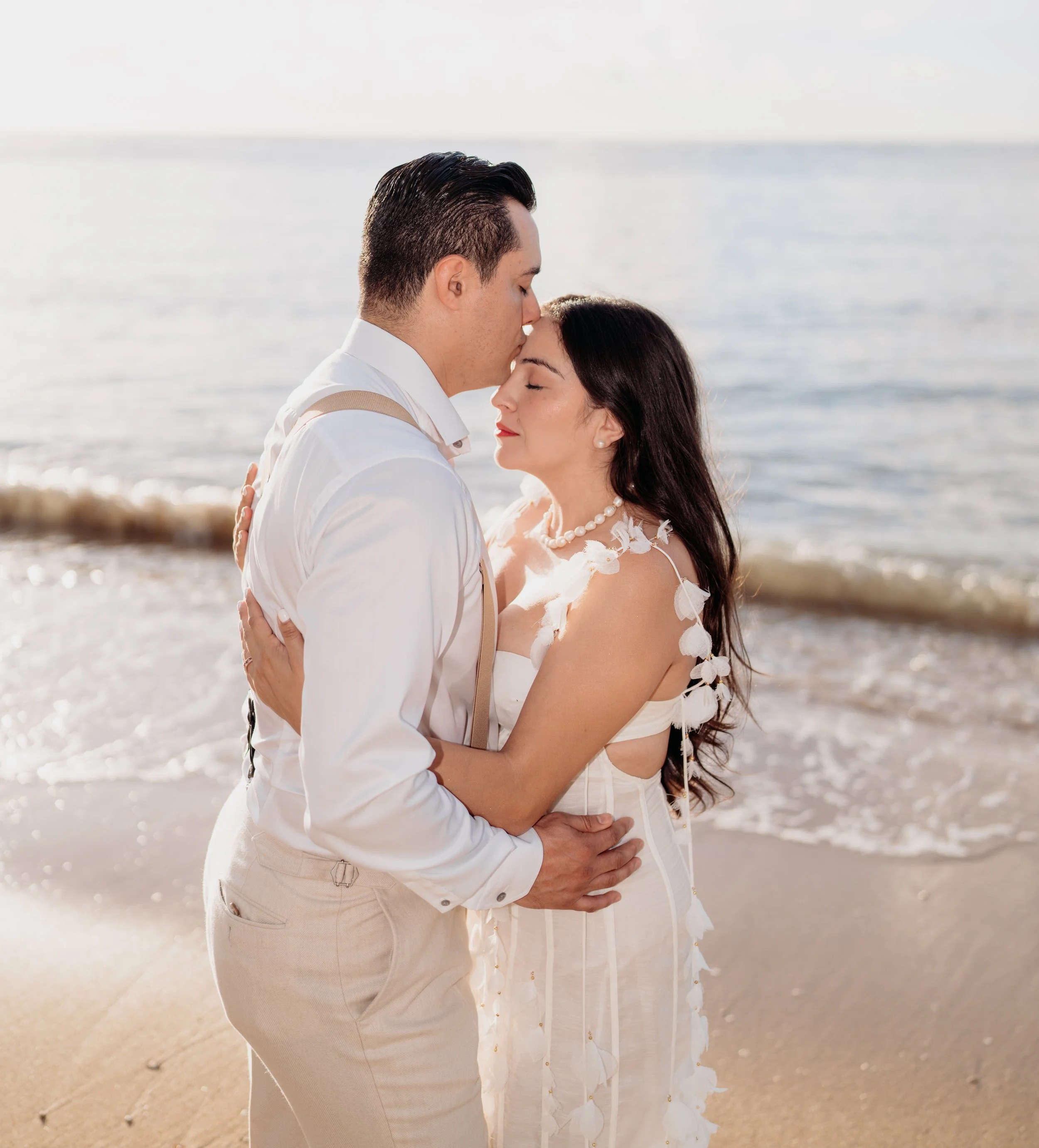 A couple embracing on a beach, with the man kissing the woman's forehead. The woman has long dark hair and is wearing a white dress with leaf details, while the man is dressed in a white shirt with suspenders. The ocean and a clear sky are in the bac