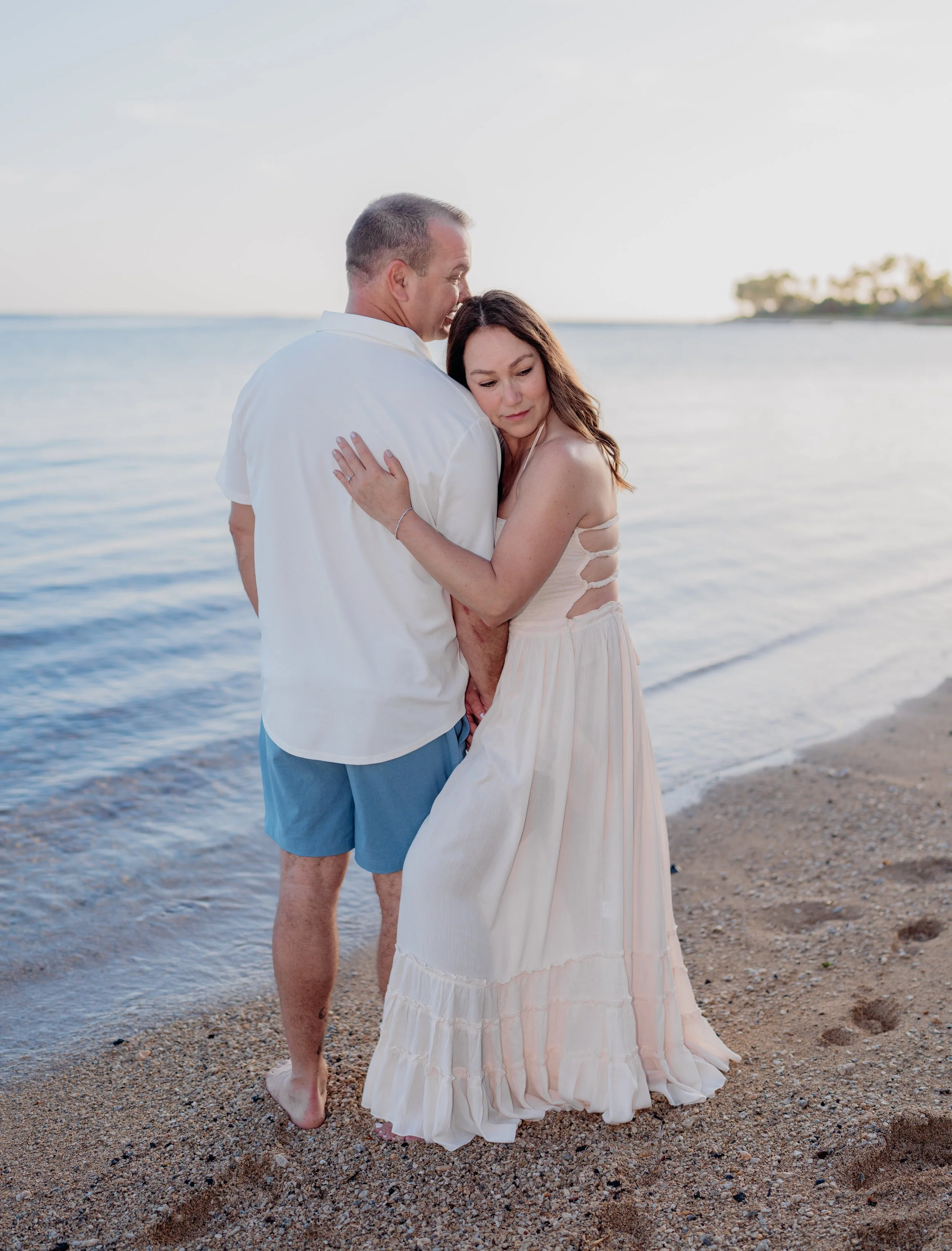 A couple hugging on a beach during sunset, dressed in casual summer clothes.