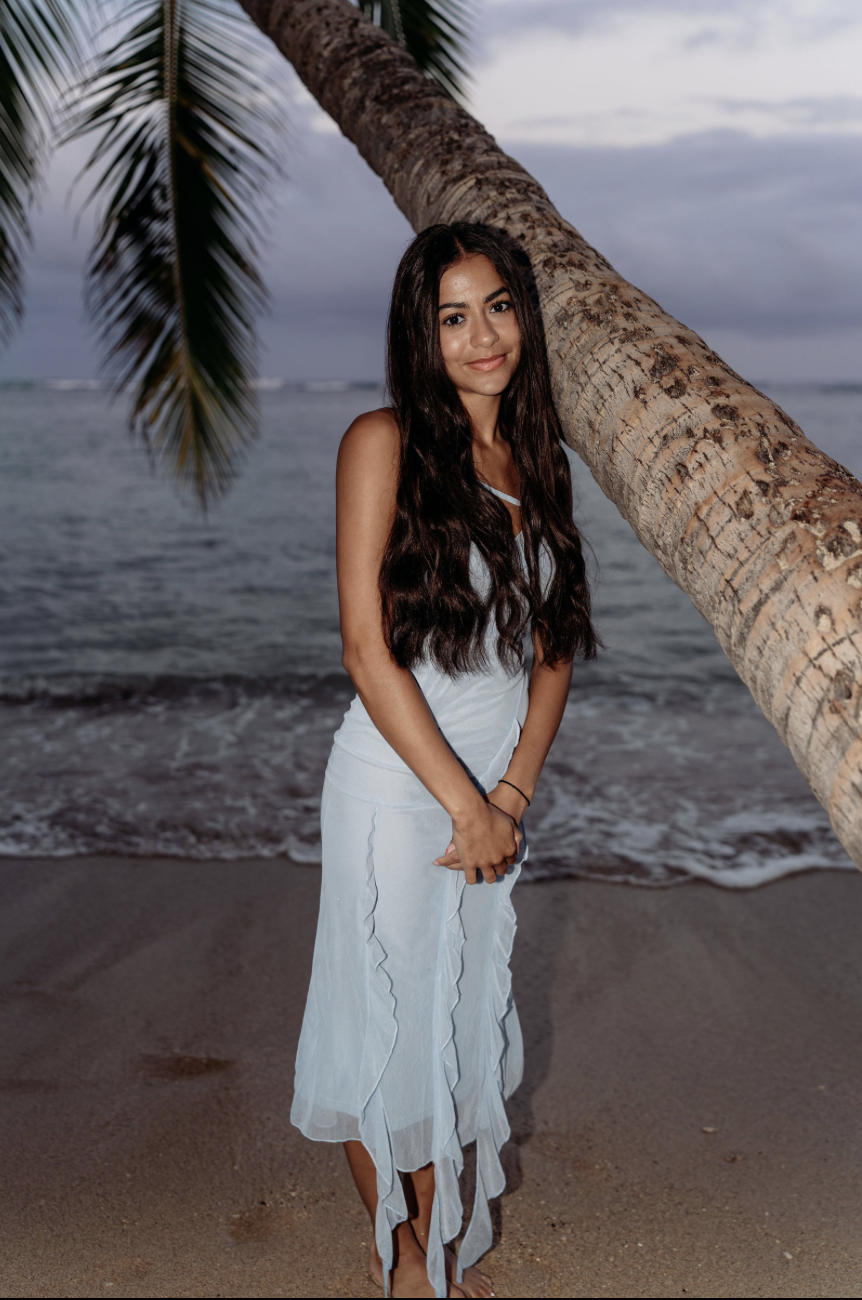 Young woman in a white dress standing on the beach near a leaning palm tree, with the ocean and a cloudy sky in the background.