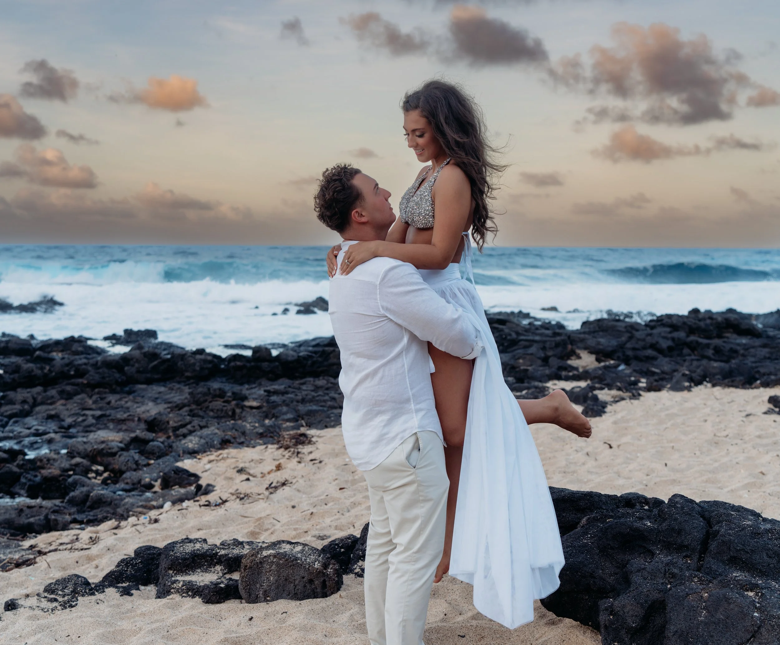 A man lifting a woman on a beach with rocks and ocean waves in the background during sunset.