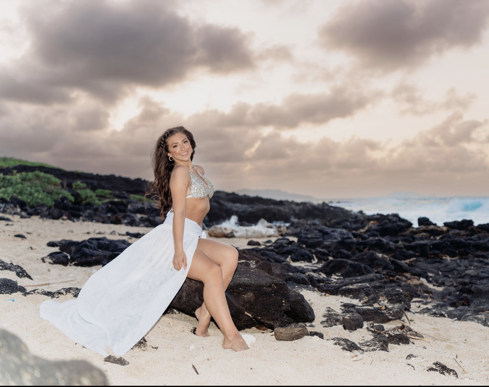 A woman in a sparkly halter top and white flowing skirt sitting on a rock on a beach with sand, rocks, ocean waves, and cloudy sky in the background.