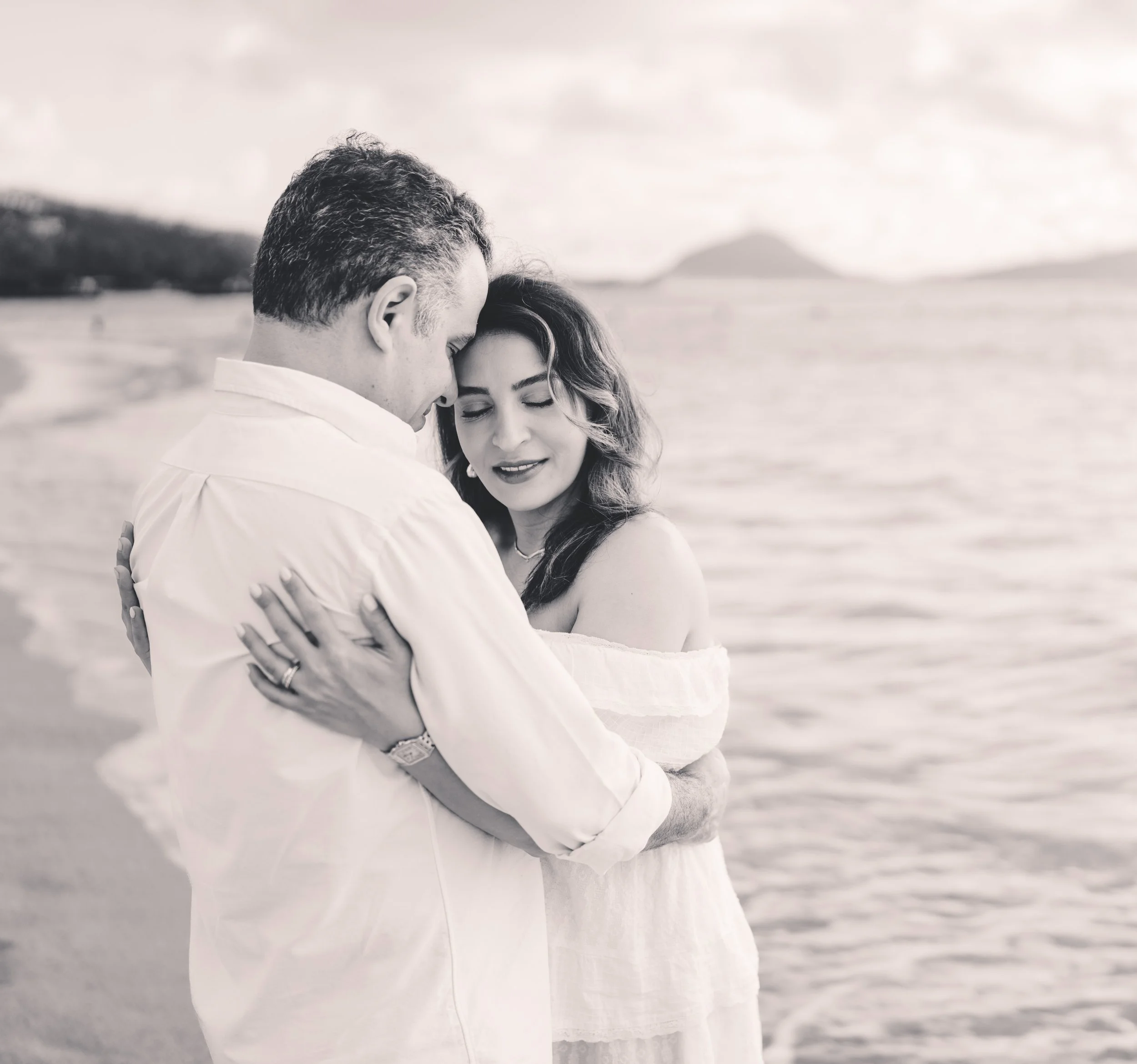 A black and white photo of a romantic moment between a man and a woman on the beach, close-eyed, embracing each other.