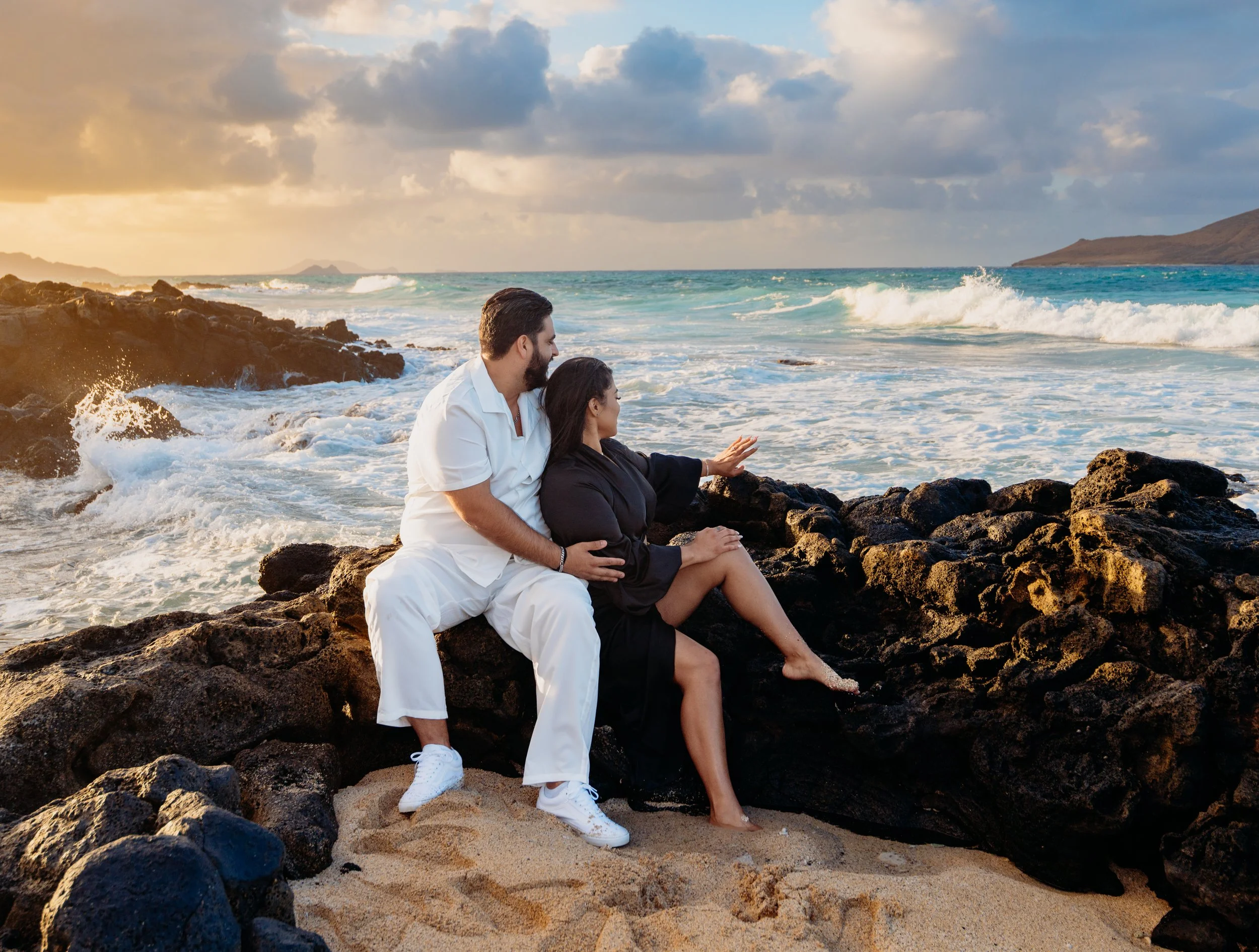 A couple sitting on rocks by the beach at sunset, with the ocean and cloudy sky in the background.