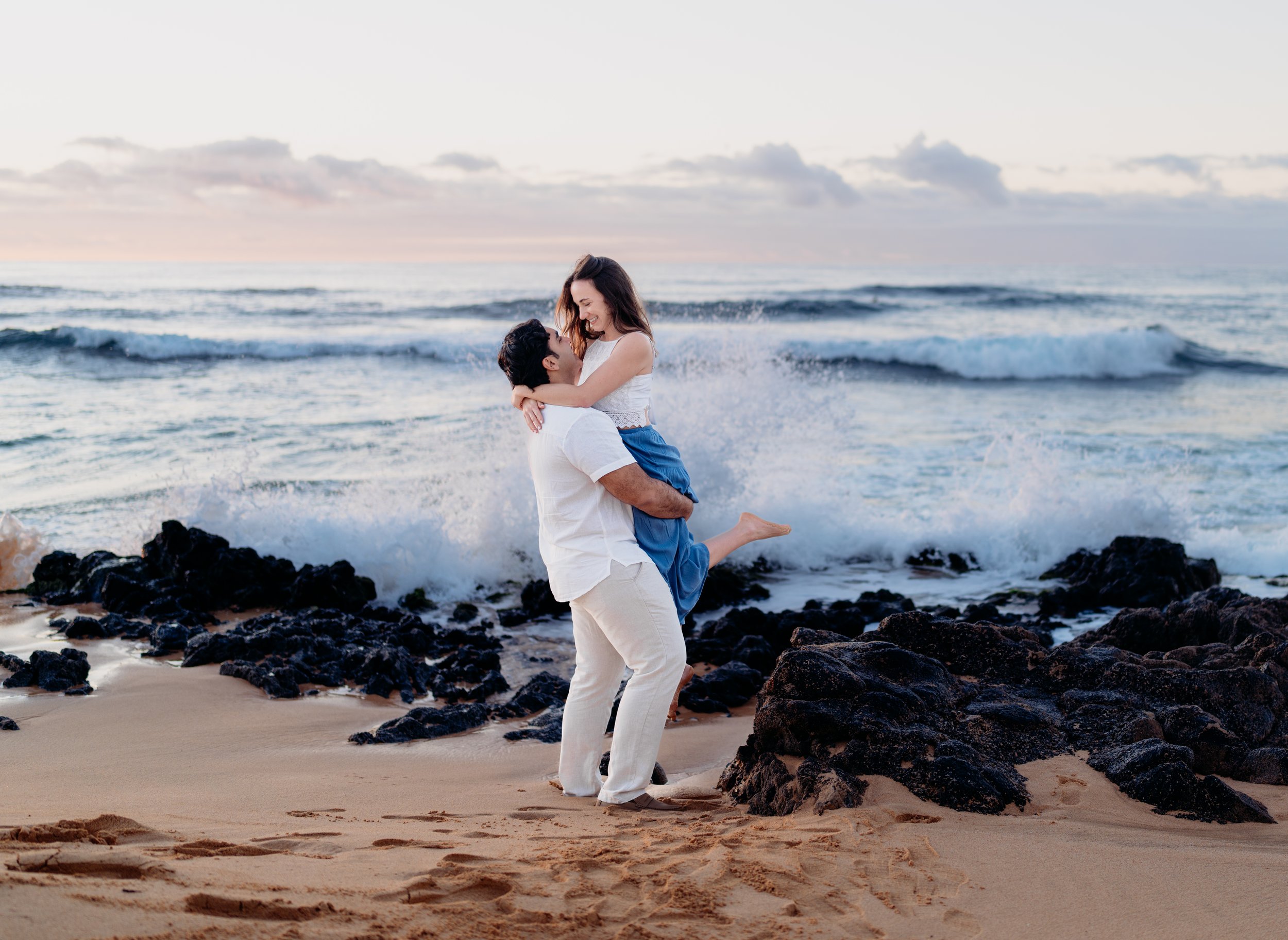 A couple on the beach, with the man lifting the woman, both smiling, near rocks and ocean waves at sunset.