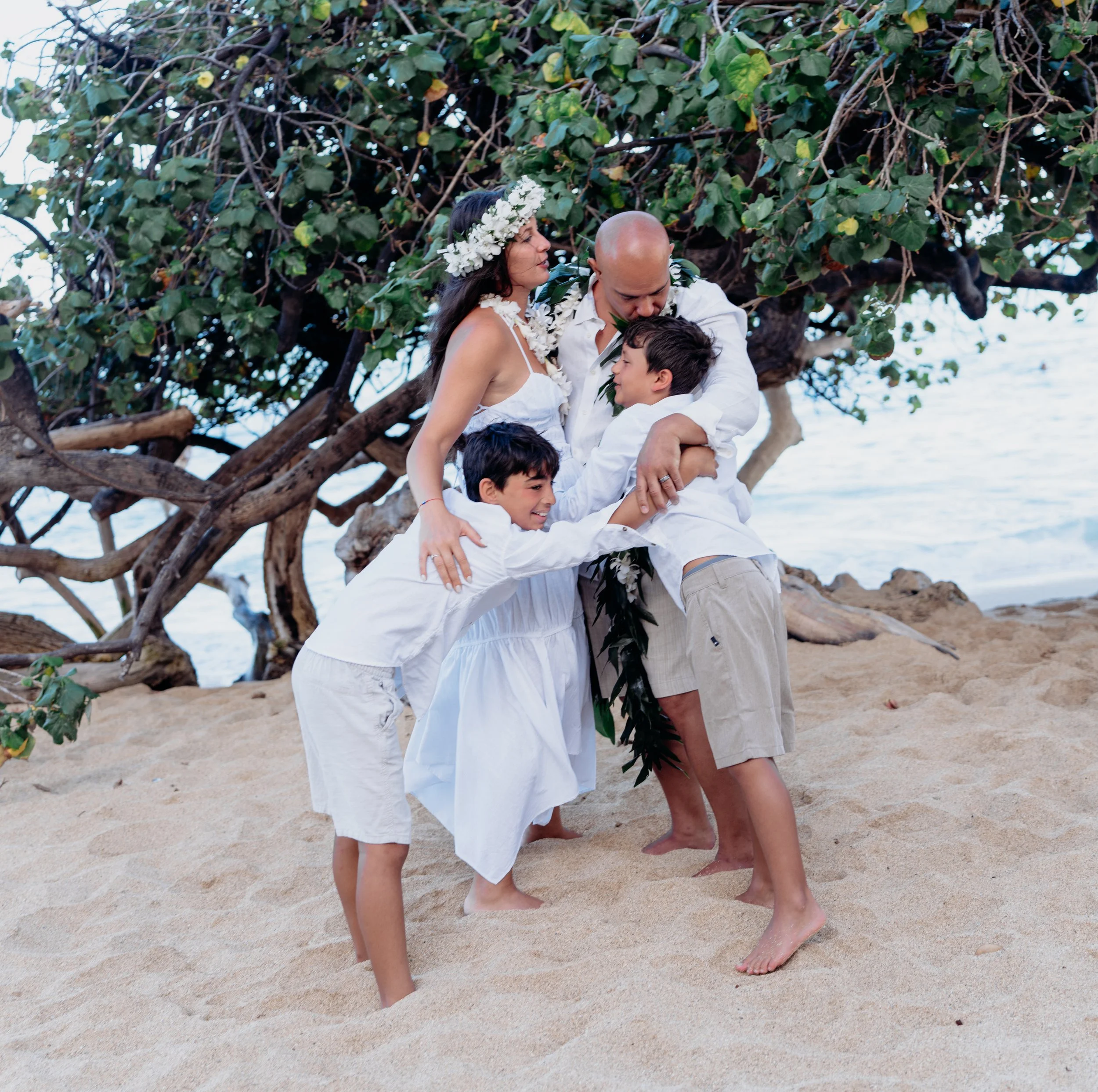 A family of four, dressed in white, hugging and playing on a sandy beach with a large tree in the background.