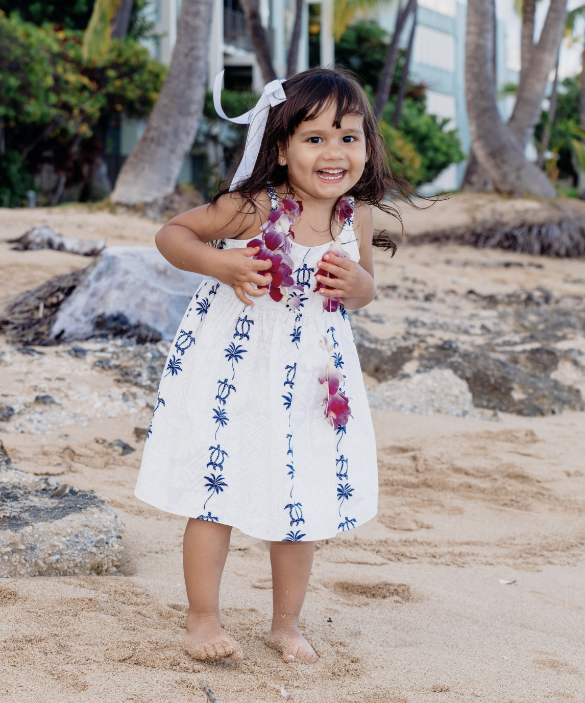 A young girl wearing a white dress with blue palm tree designs, a white hair bow, and a lei, smiling and standing barefoot on a sandy beach with trees and buildings in the background.