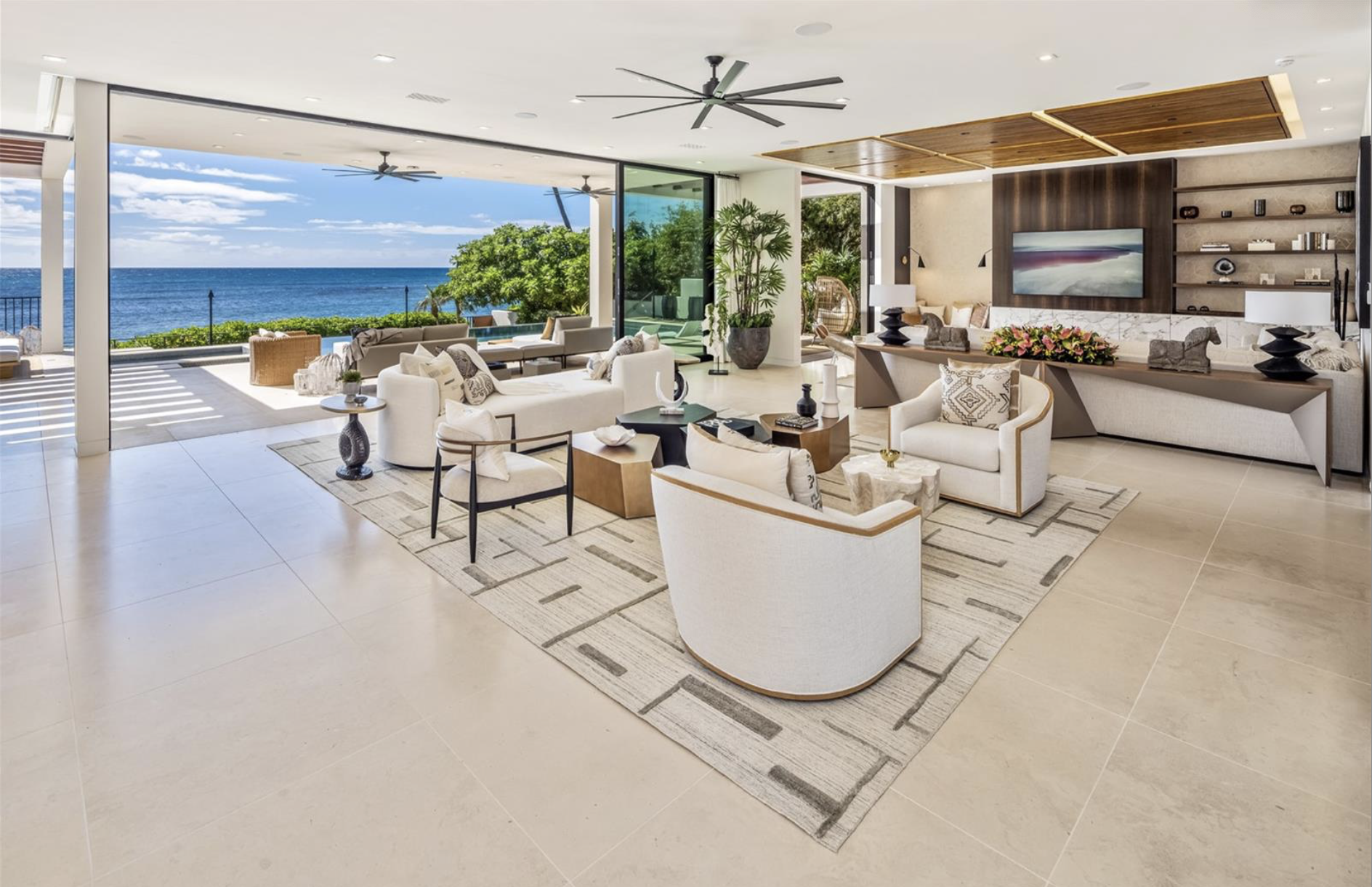 Modern living room with large glass sliding doors opening to ocean view, featuring white sofas, armchairs, a patterned rug, decorative plants, and a flat-screen TV on a dark wood accent wall.