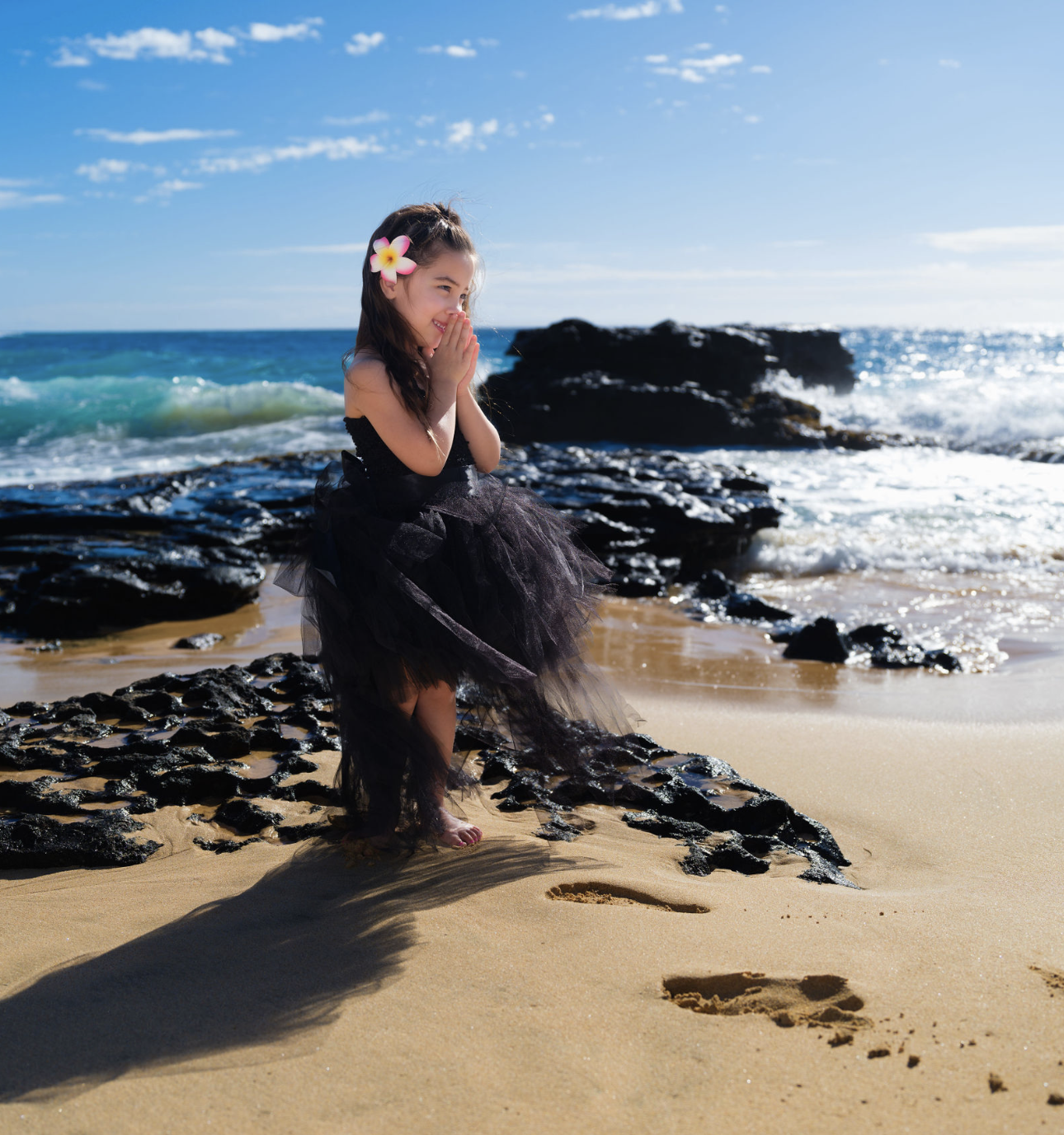 A young girl in a black tulle dress and a pink flower in her hair stands on the beach, smiling and holding her hands near her face, with footprints in the sand behind her and the ocean and rocks in the background.