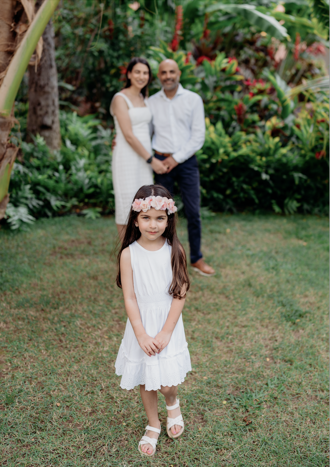 Young girl in a white dress and pink floral headband standing outdoors on grass, with a man and woman in white outfits smiling and standing among lush greenery in the background.