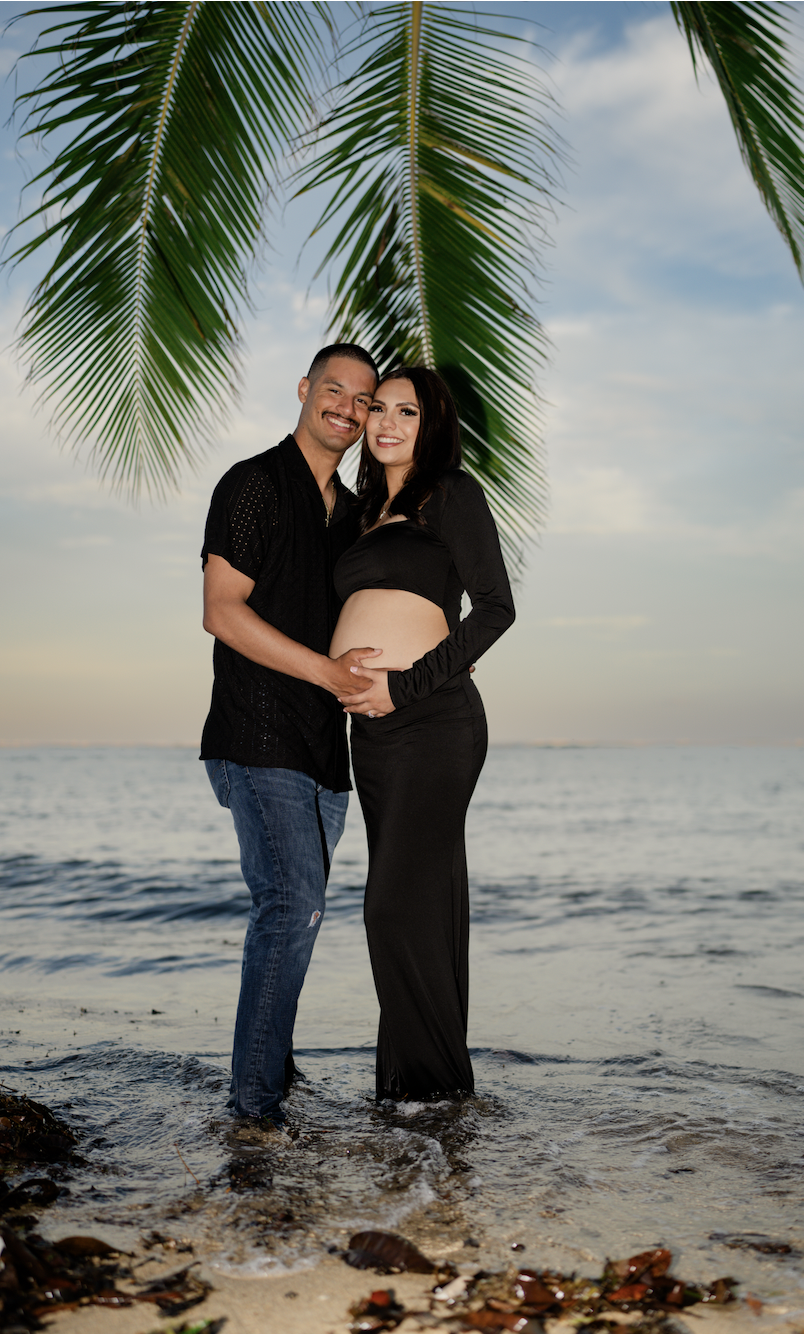 A happy couple on the beach during sunset, with palm tree leaves overhead. The woman is pregnant and wearing a black crop top and skirt, and the man is dressed in a black shirt and jeans. They are smiling and holding her belly, standing in shallow water.