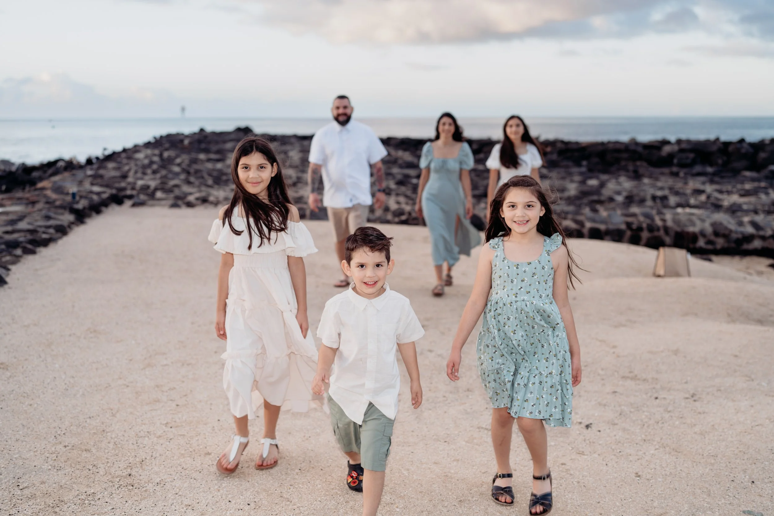 A family of six walking along a sandy path near the sea during sunset, with rocky formations in the background and clouds in the sky.