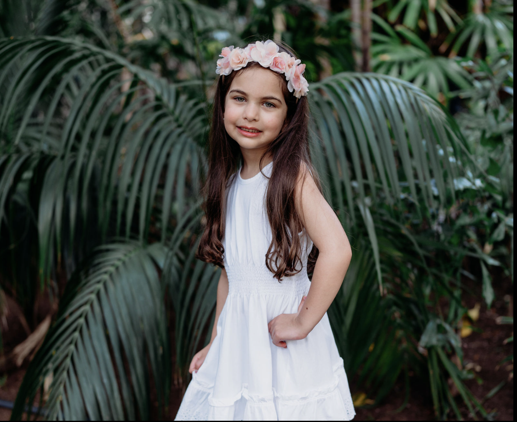 Young girl with long brown hair wearing a white dress and a pink flower crown, standing outdoors among green tropical plants.