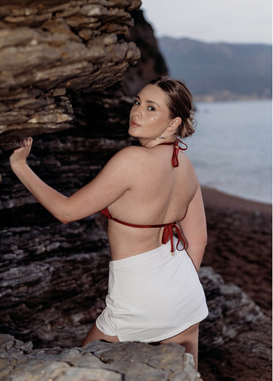A woman with light skin and brunette hair styled in a bun, wearing a red bikini top and white skirt, standing by rocky terrain near a body of water with mountains in the background, looking over her shoulder at the camera.