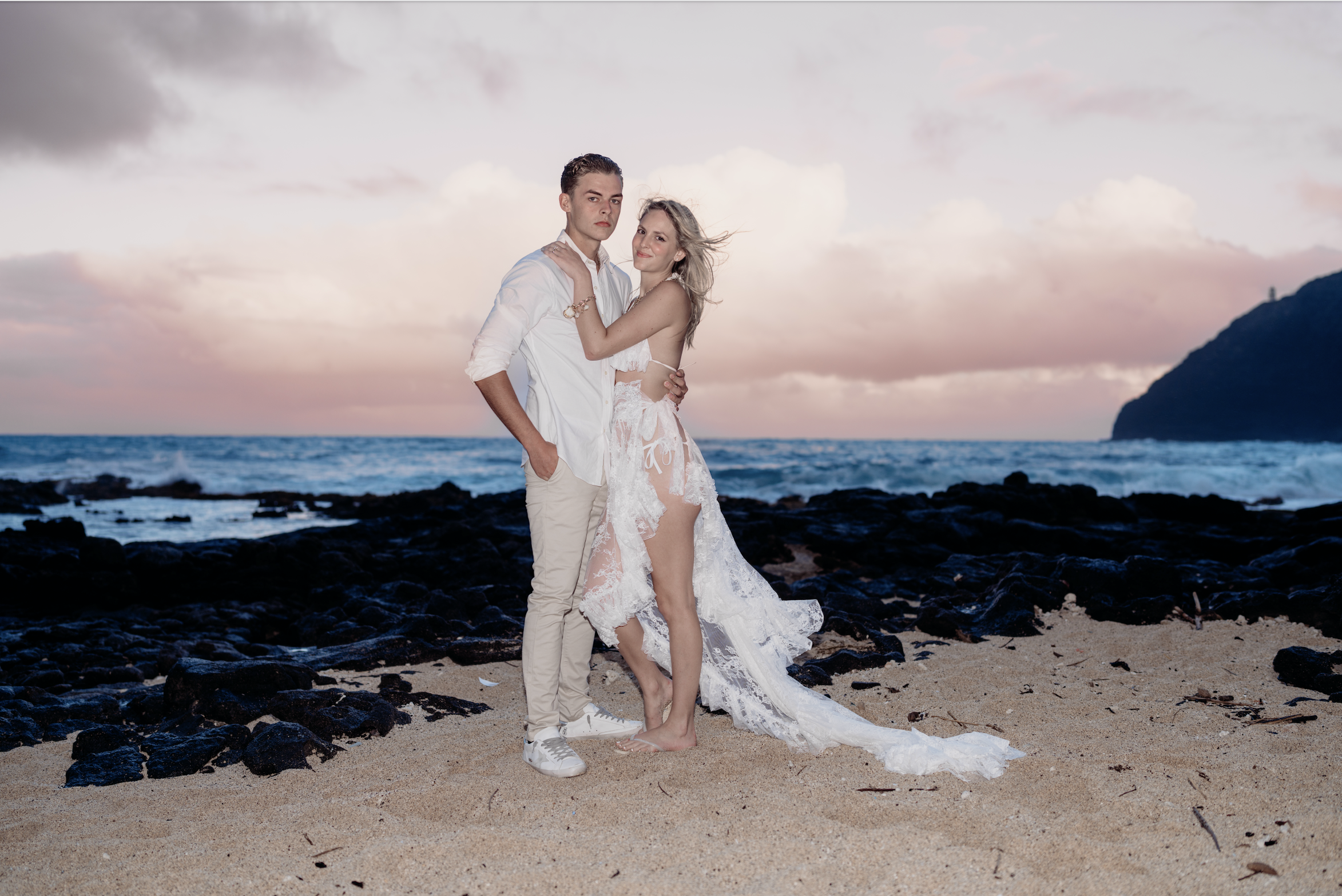 A young couple in wedding attire on a beach at sunset, with the ocean and a rocky coastline in the background.