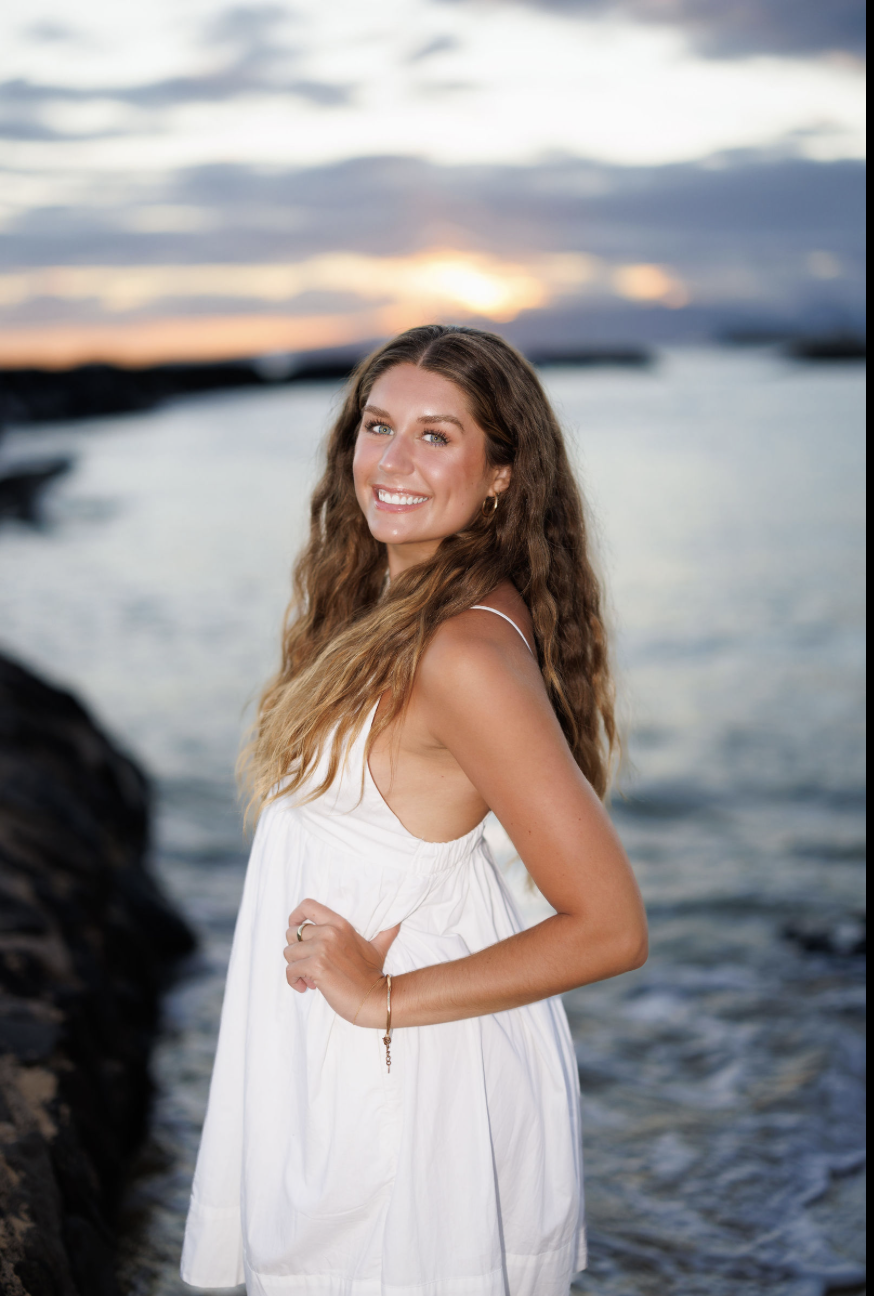 A woman with long wavy brown hair smiling at the camera, standing on a beach with rocks and water in the background during sunset, wearing a white sleeveless dress.
