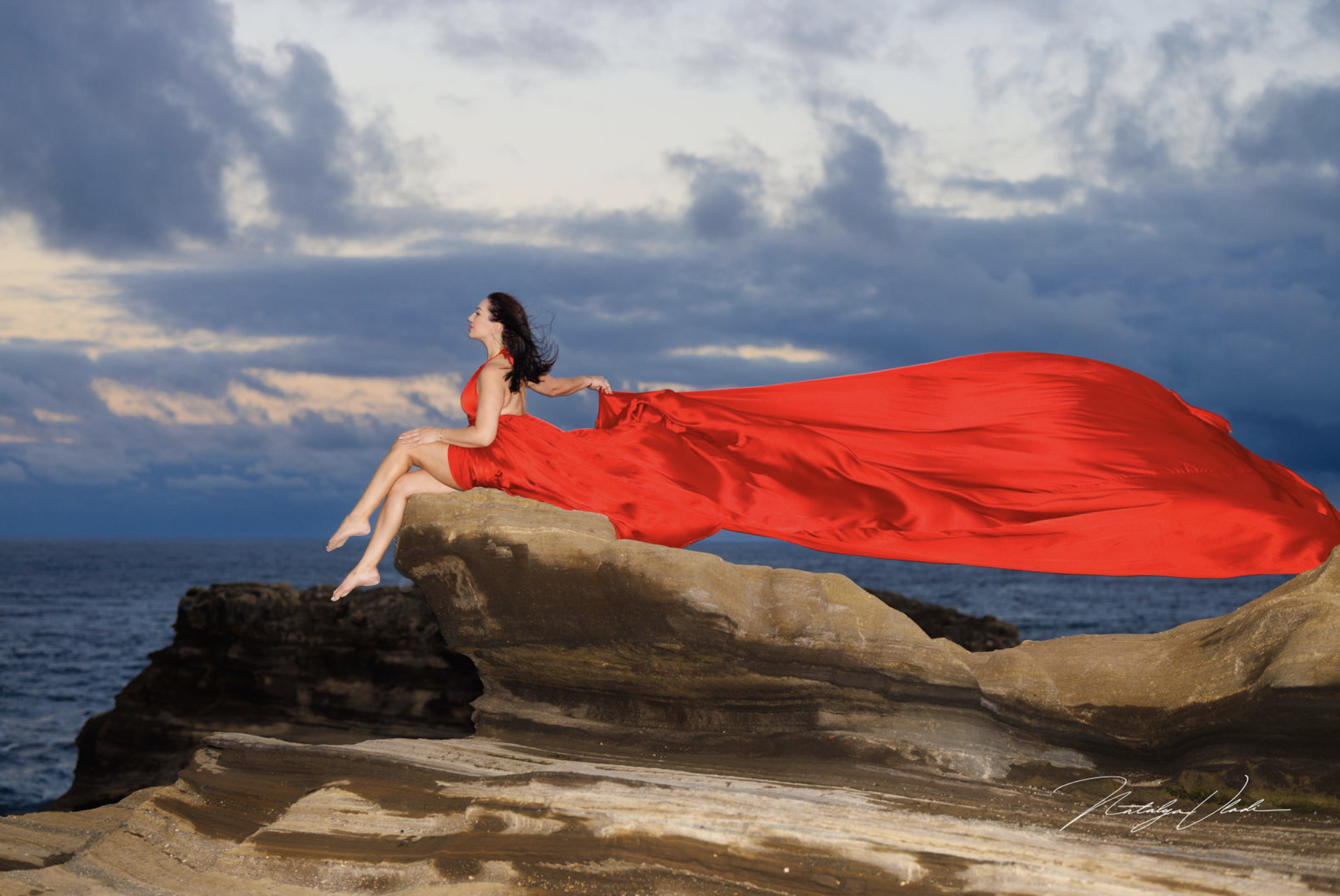 A woman in a flowing red dress sitting on a rock by the ocean, with a cloudy sky in the background.