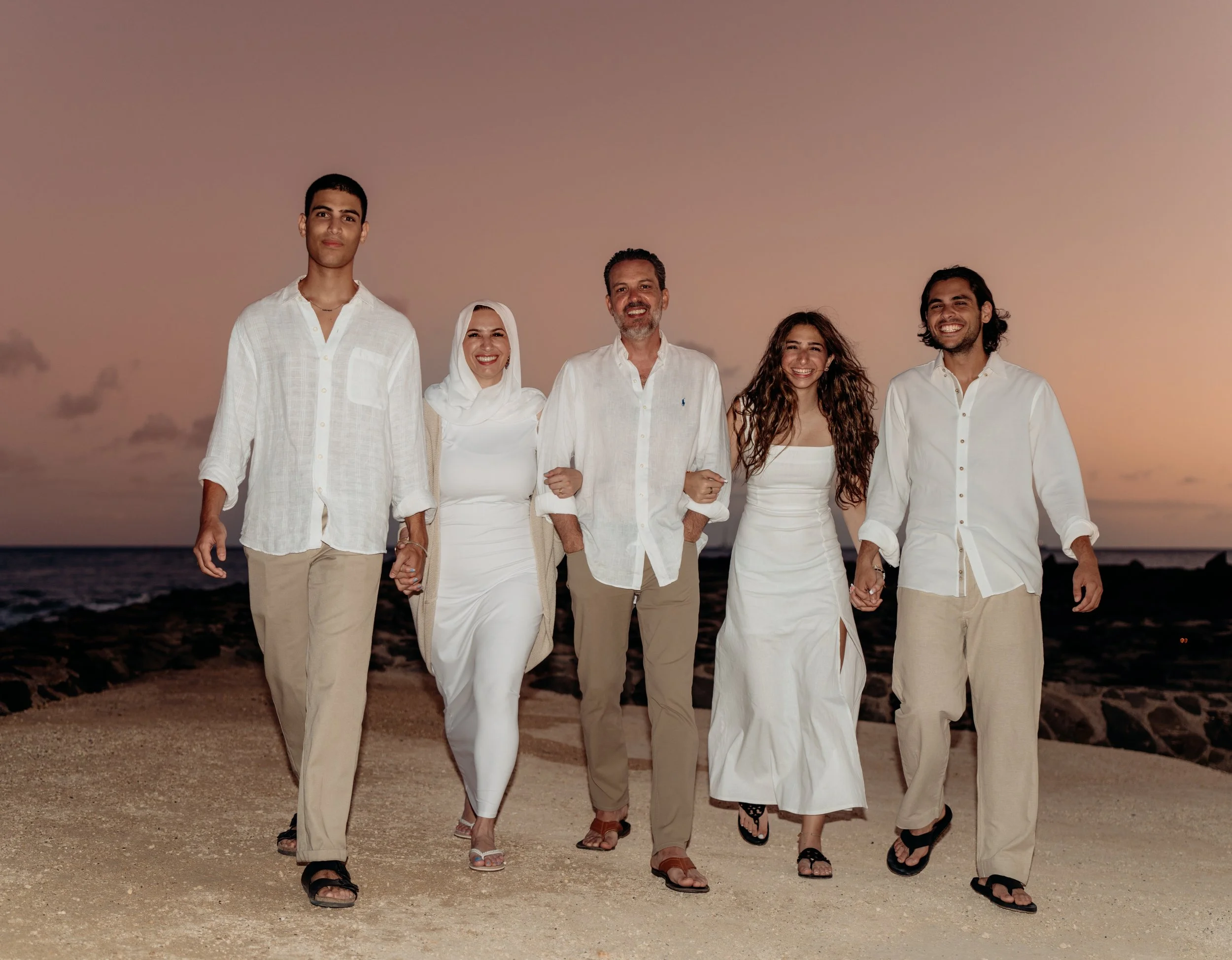 Group of five people walking on a beach at sunset, holding hands and smiling, dressed in white and beige casual summer clothing.