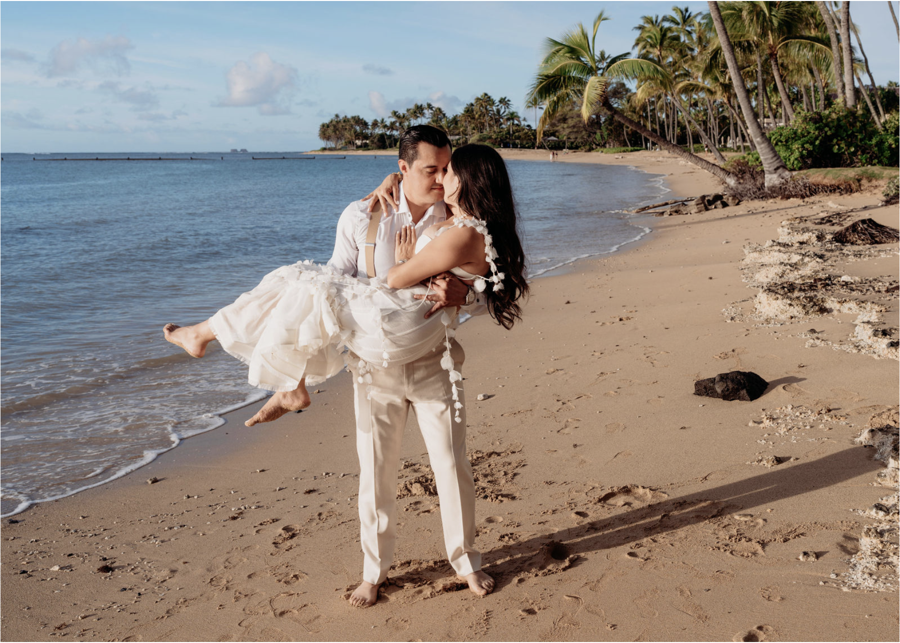 A man in white pants and shirt holding a woman in a white dress with floral details on a sandy beach with palm trees and ocean in the background.