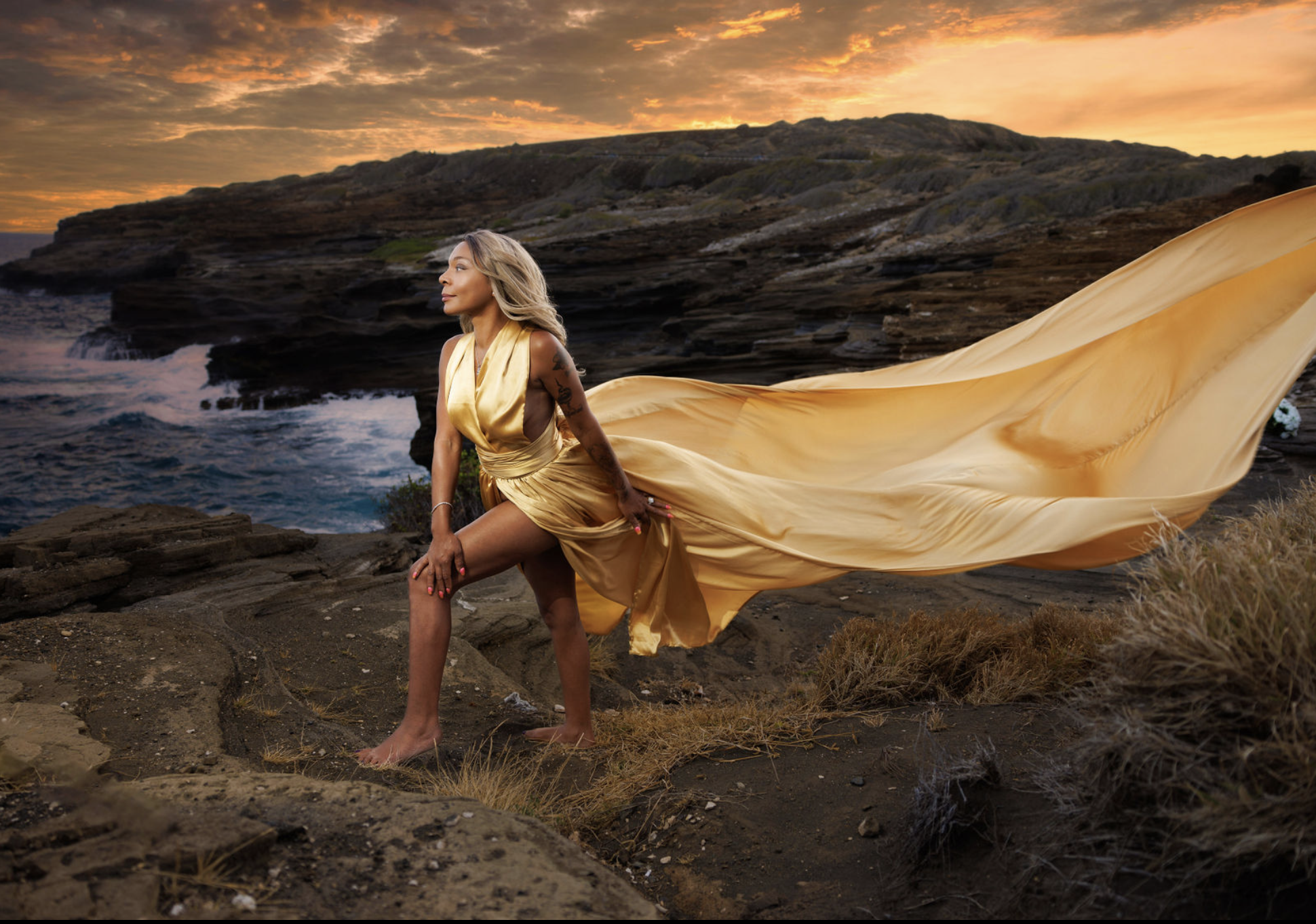 A woman in a flowing gold dress stands barefoot on rocky terrain by the ocean at sunset, with cliffs in the background.