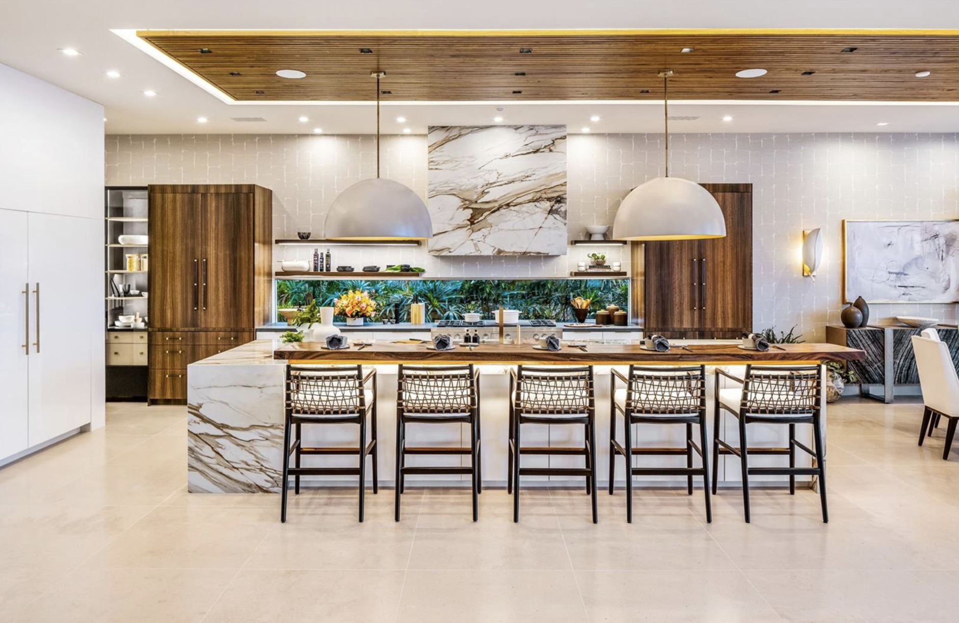 Modern kitchen with a marble island, six chairs, wooden cabinets, a marble artwork, two large pendant lights, a backsplash with greenery, and decorative items.