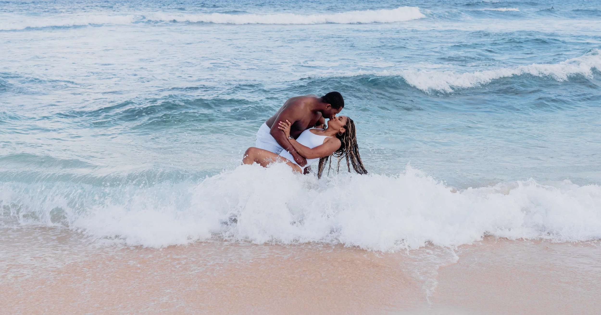 A couple, a man and a woman, are in the ocean at the beach, with the man holding the woman as they kiss, waves in the background.