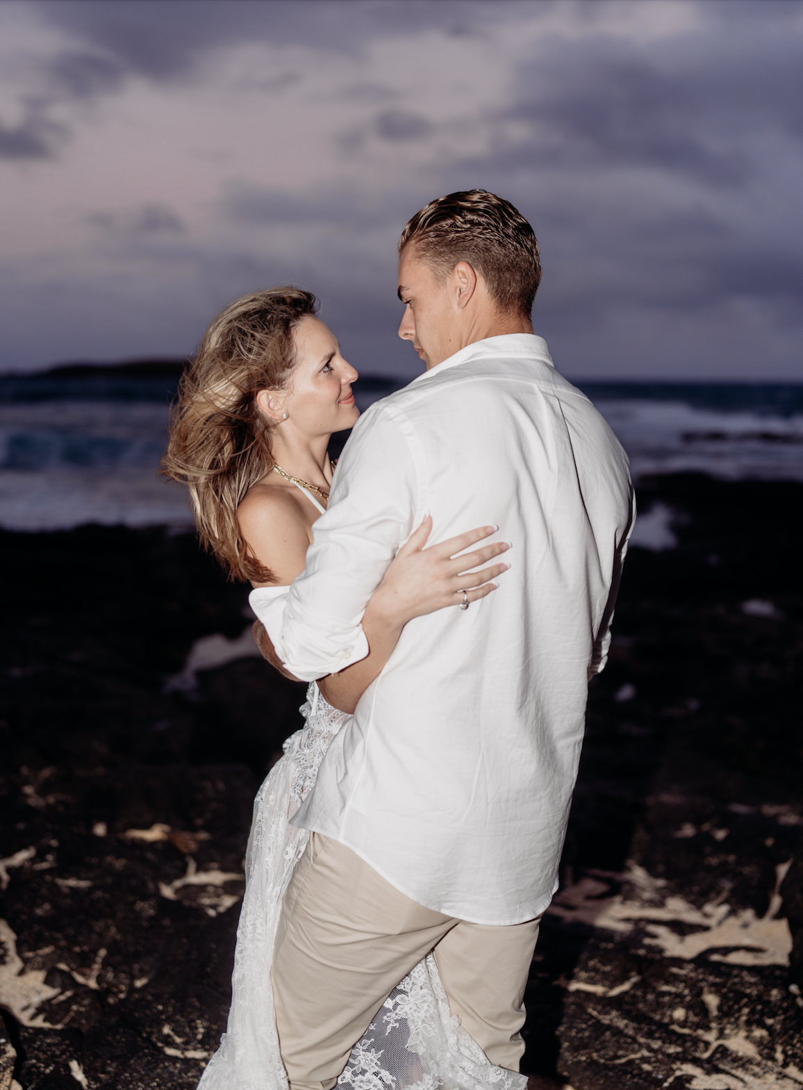 A couple dancing on a beach during sunset, with the ocean and cloudy sky in the background.