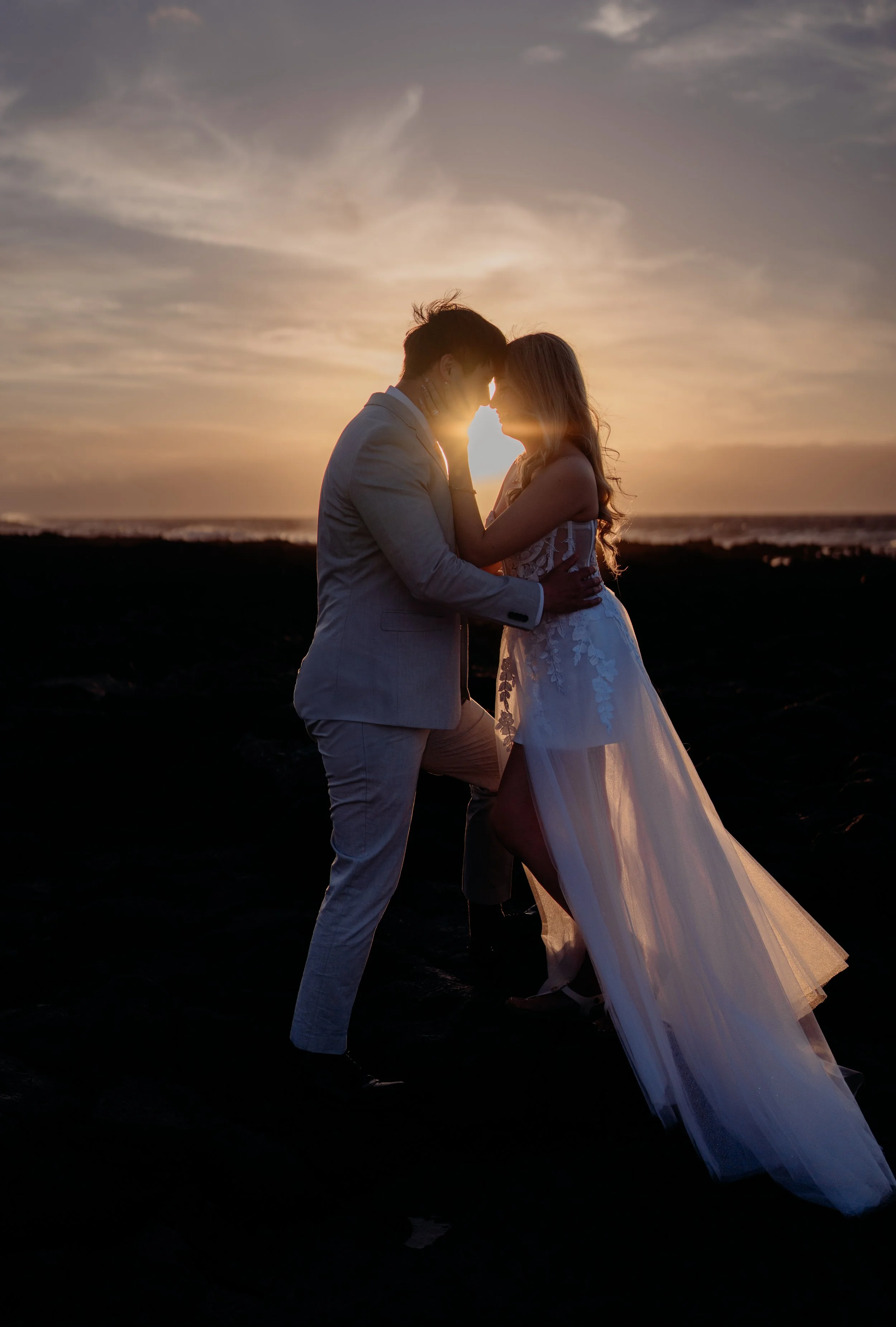 A couple dressed in wedding attire couple are standing close to each other on a beach during sunset, with their foreheads touching and backlit by the setting sun.