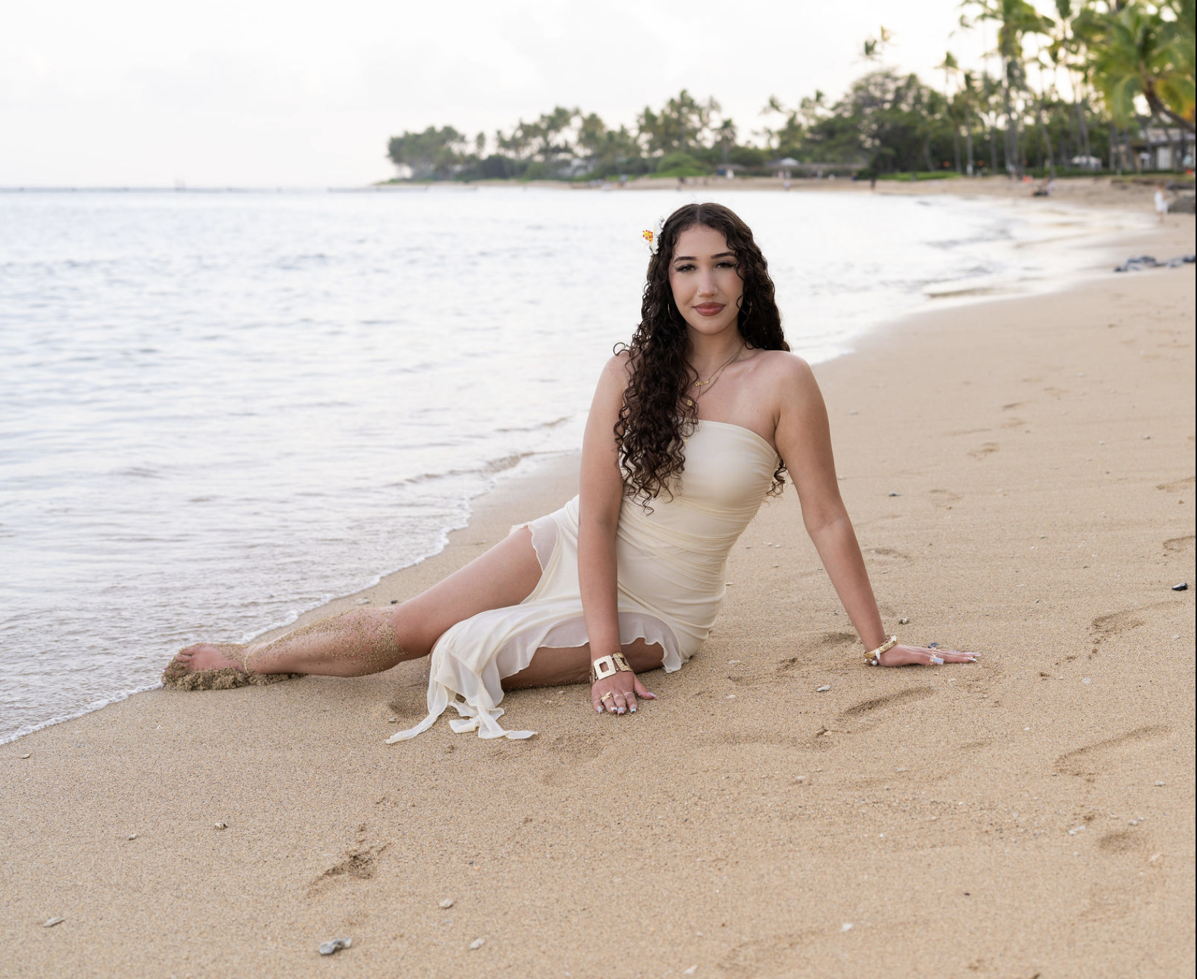 A woman in a cream-colored dress sitting on a sandy beach near the water, with trees and buildings in the background.